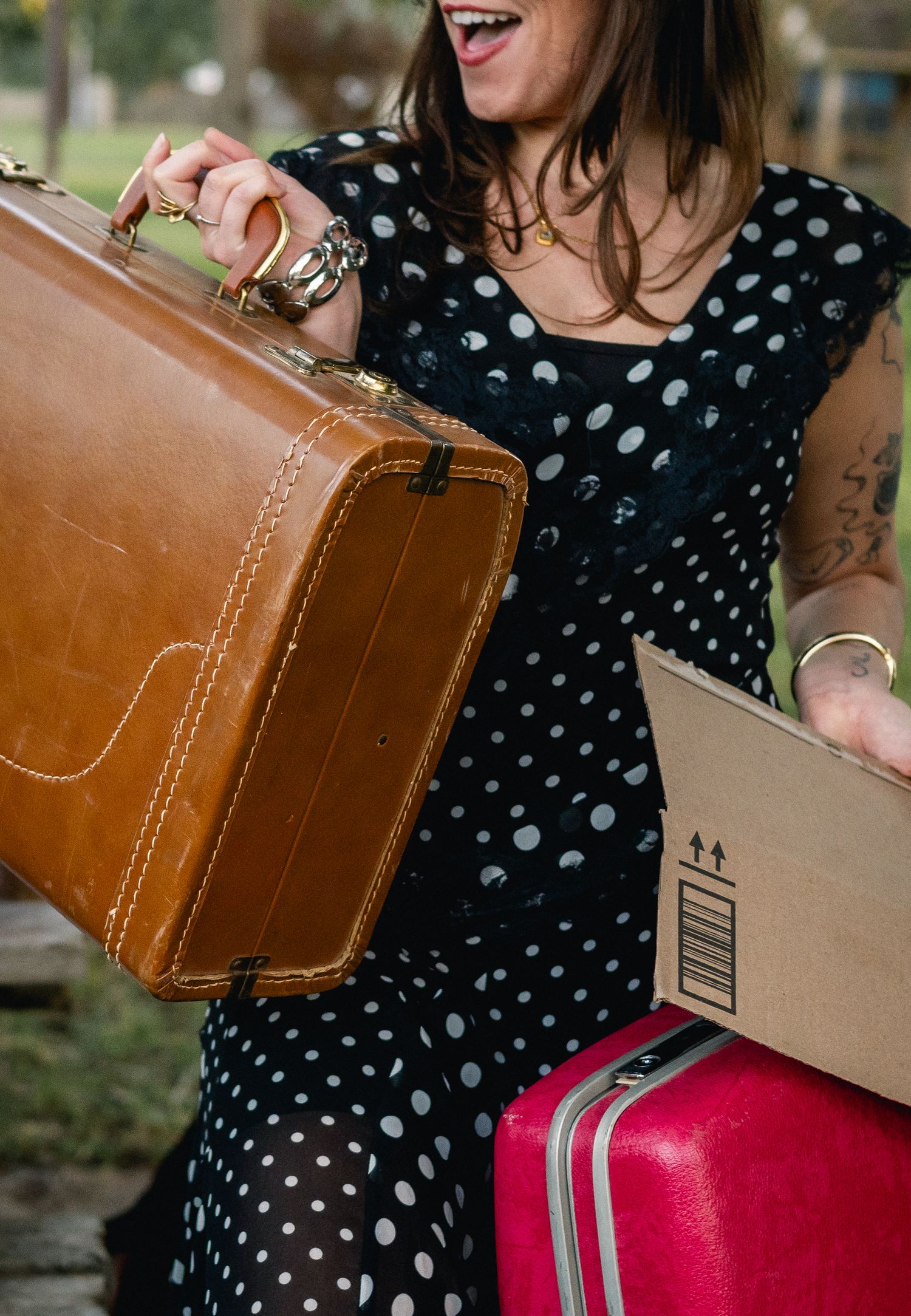 Woman in polka dot dress carrying brown vintage suitcase, cardboard box, and pink luggage while smiling.