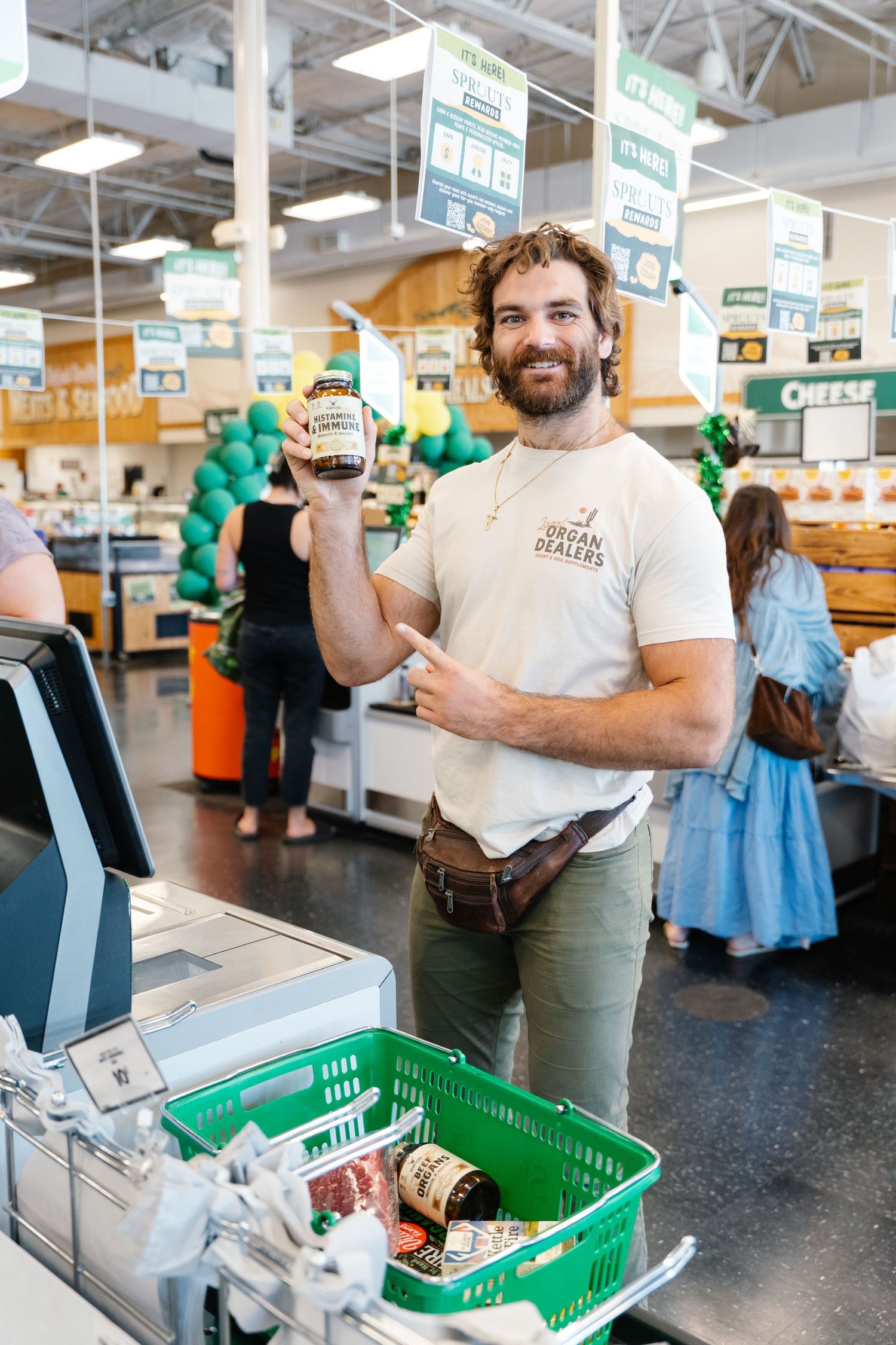 Smiling bearded man in organic dealers shirt holds up a product jar at a grocery store checkout.