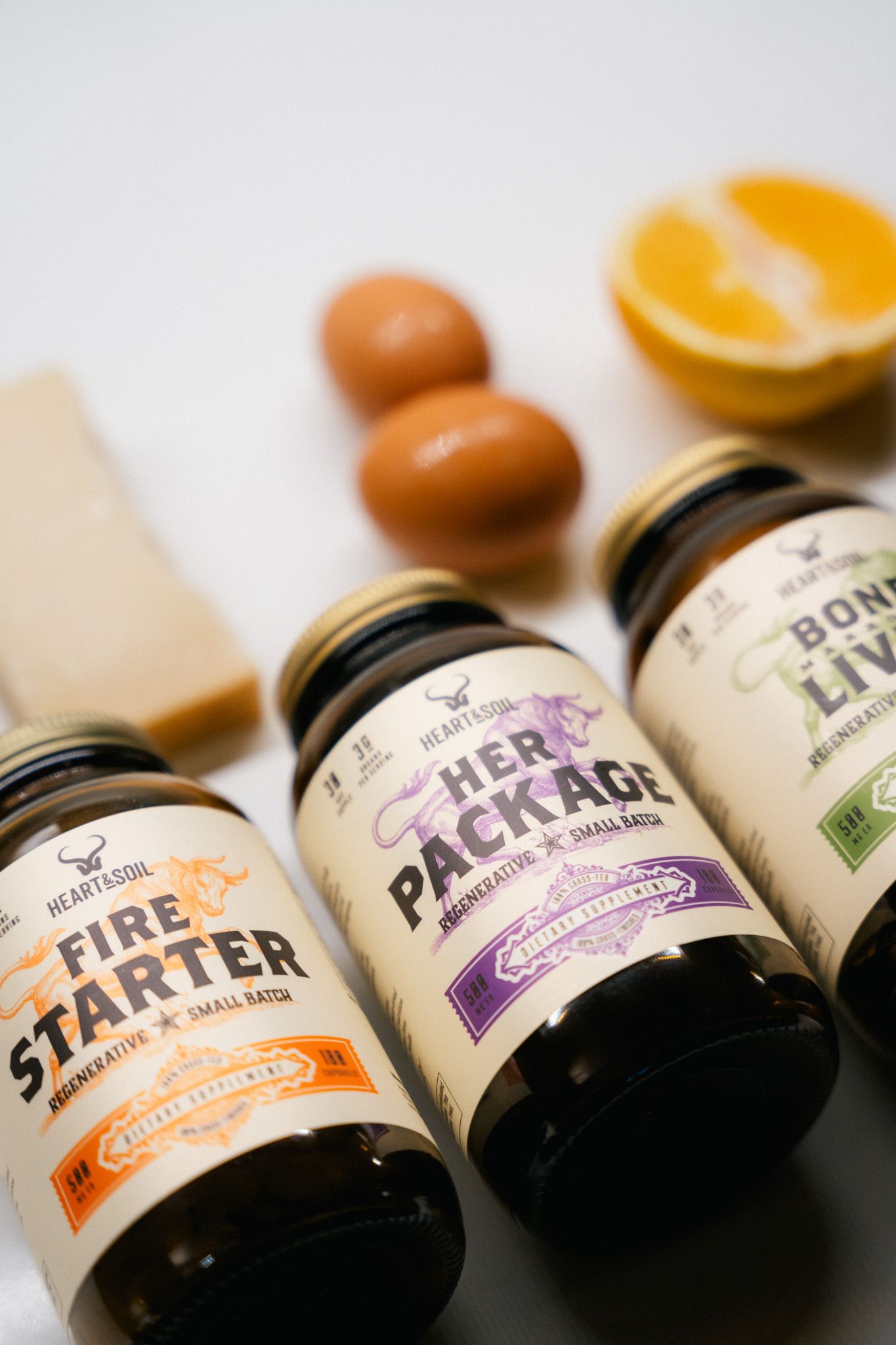 Three Heart & Soil supplement jars labeled Fire Starter, Her Package, and Bone & Liver displayed with eggs and orange.