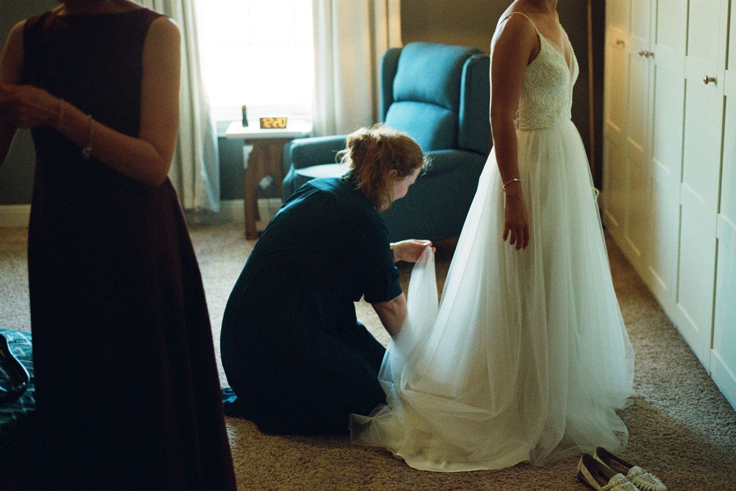 Wedding assistant kneels to adjust bride's white tulle gown hem in a softly lit room.