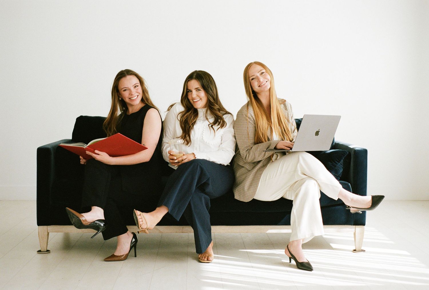Three professional women sitting on a dark sofa with a laptop and red binder in bright studio.