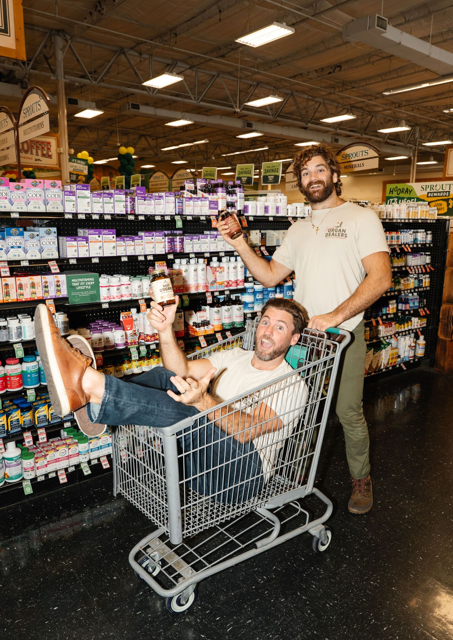 Two men posing playfully with supplements in a shopping cart inside a health food store.