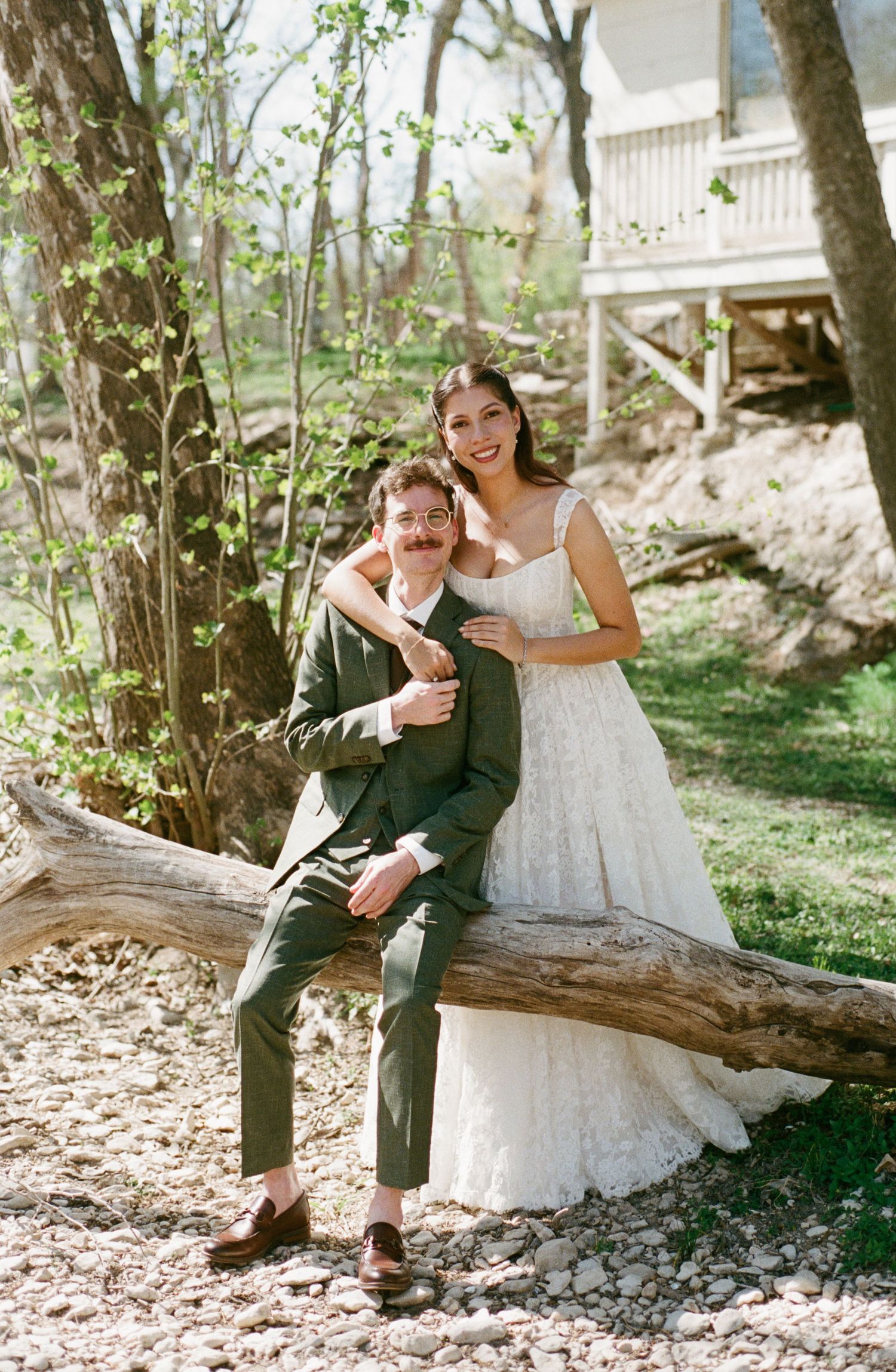 Smiling bride and groom posing on a fallen log in a wooded outdoor wedding setting in spring.