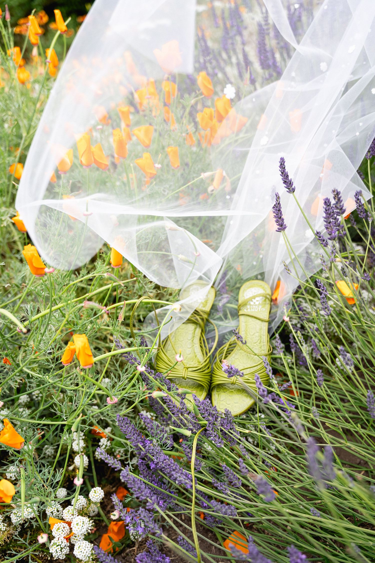 Green bridal sandals nestled among lavender and orange poppy wildflowers with a flowing white wedding veil.