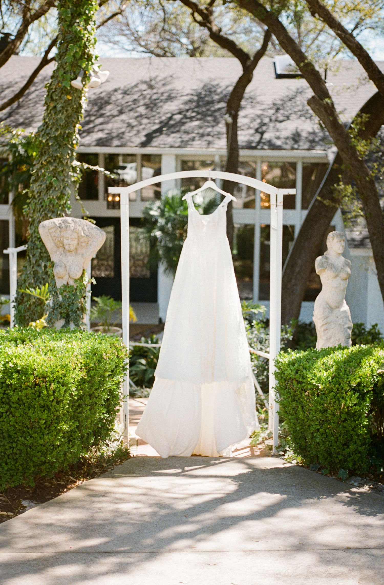 Elegant white wedding dress hanging on arch surrounded by sculpted statues and lush greenery in garden.