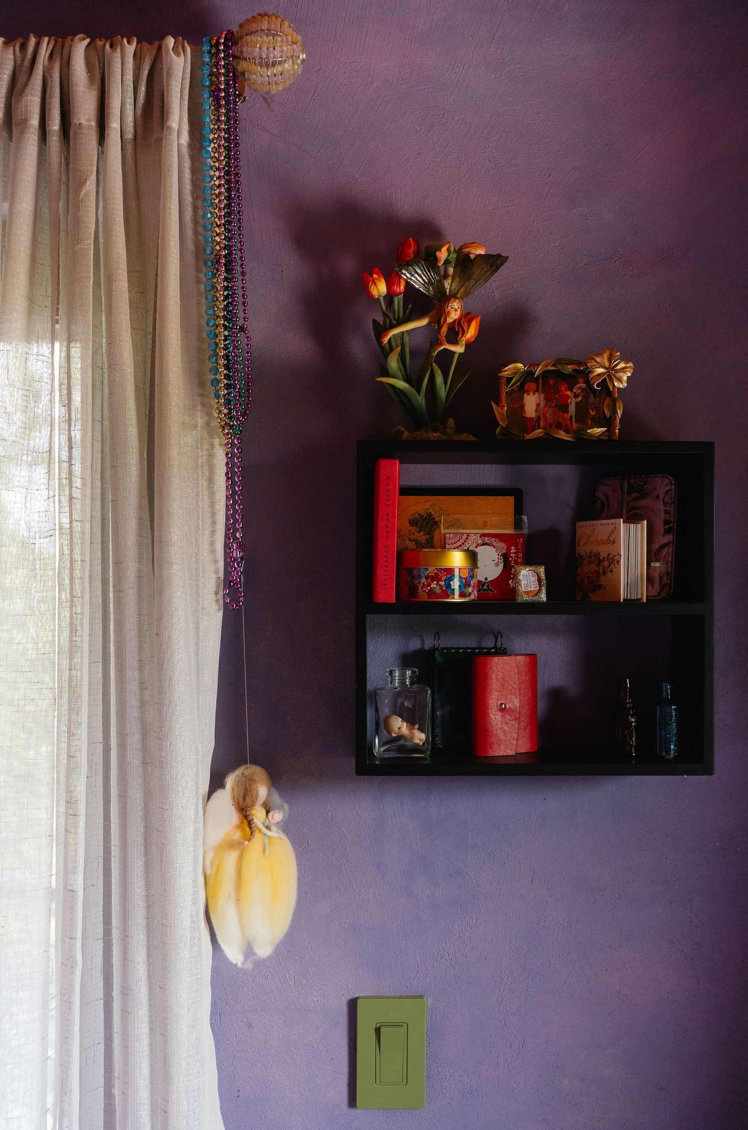 Purple wall with black shelf displaying books, plants, and decorative items beside sheer curtains with colorful beaded garland.