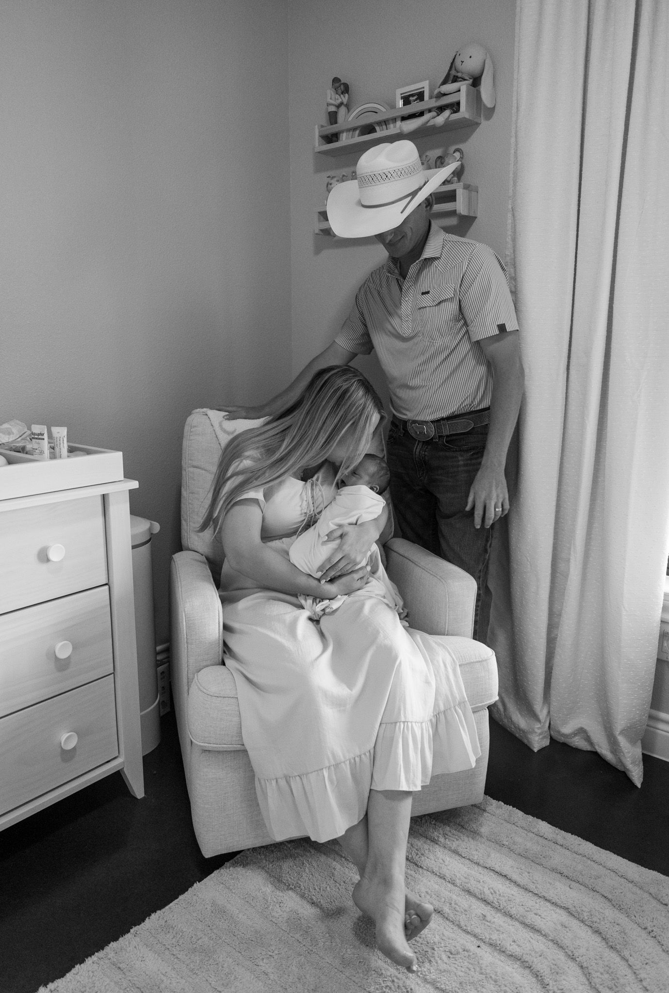 Black and white photo of new parents in nursery, mother seated holding newborn while father leans in.