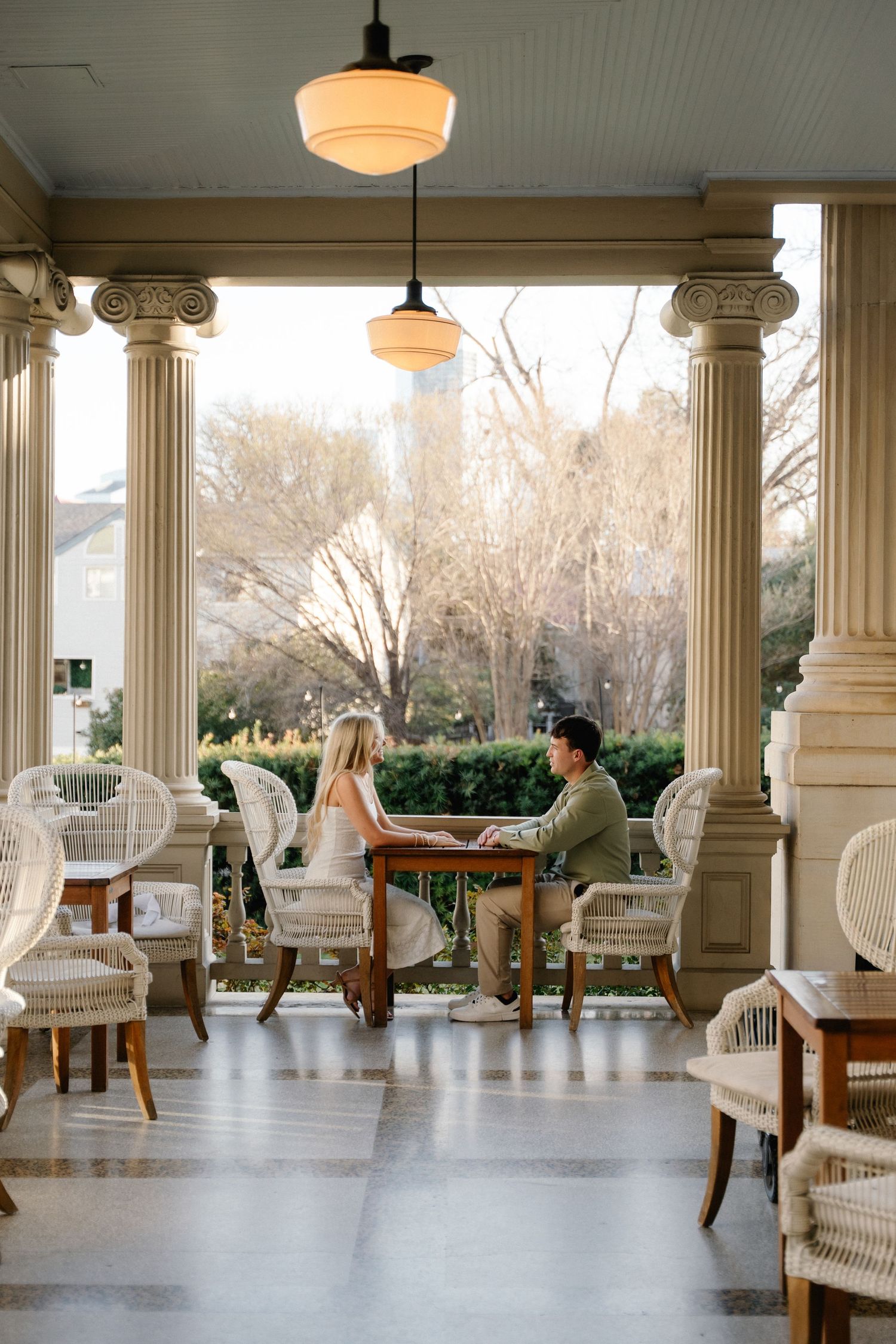 A couple sits at a wooden table on a grand columned porch with wicker chairs and warm lighting.