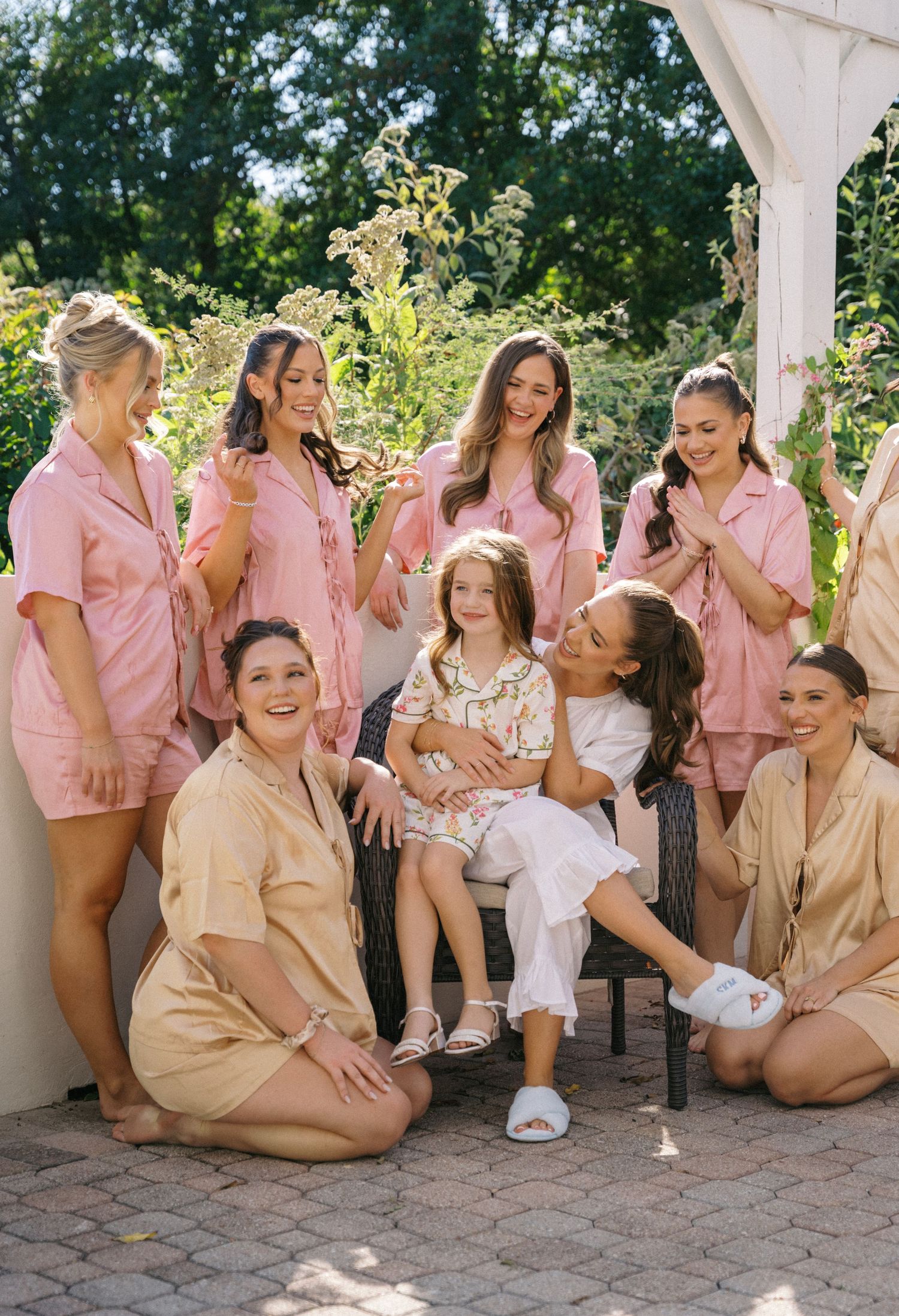 Bridesmaids in pink satin pajamas gathered outdoors with a flower girl on a sunny wedding morning.