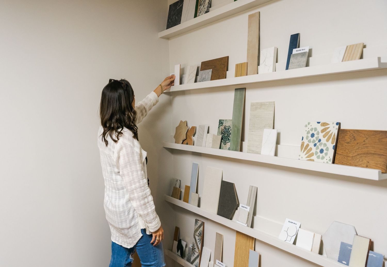 Woman browsing material sample shelves displaying tiles, fabrics, and design swatches in a showroom.