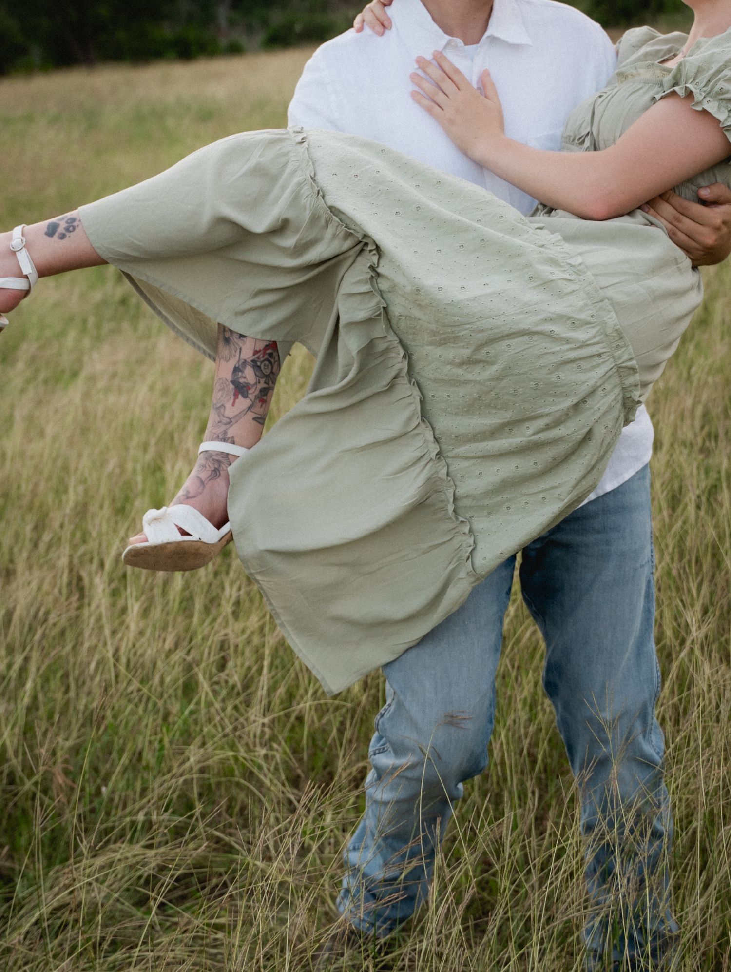 A man carries a woman in a sage green dress through a grassy field, her tattooed legs visible.