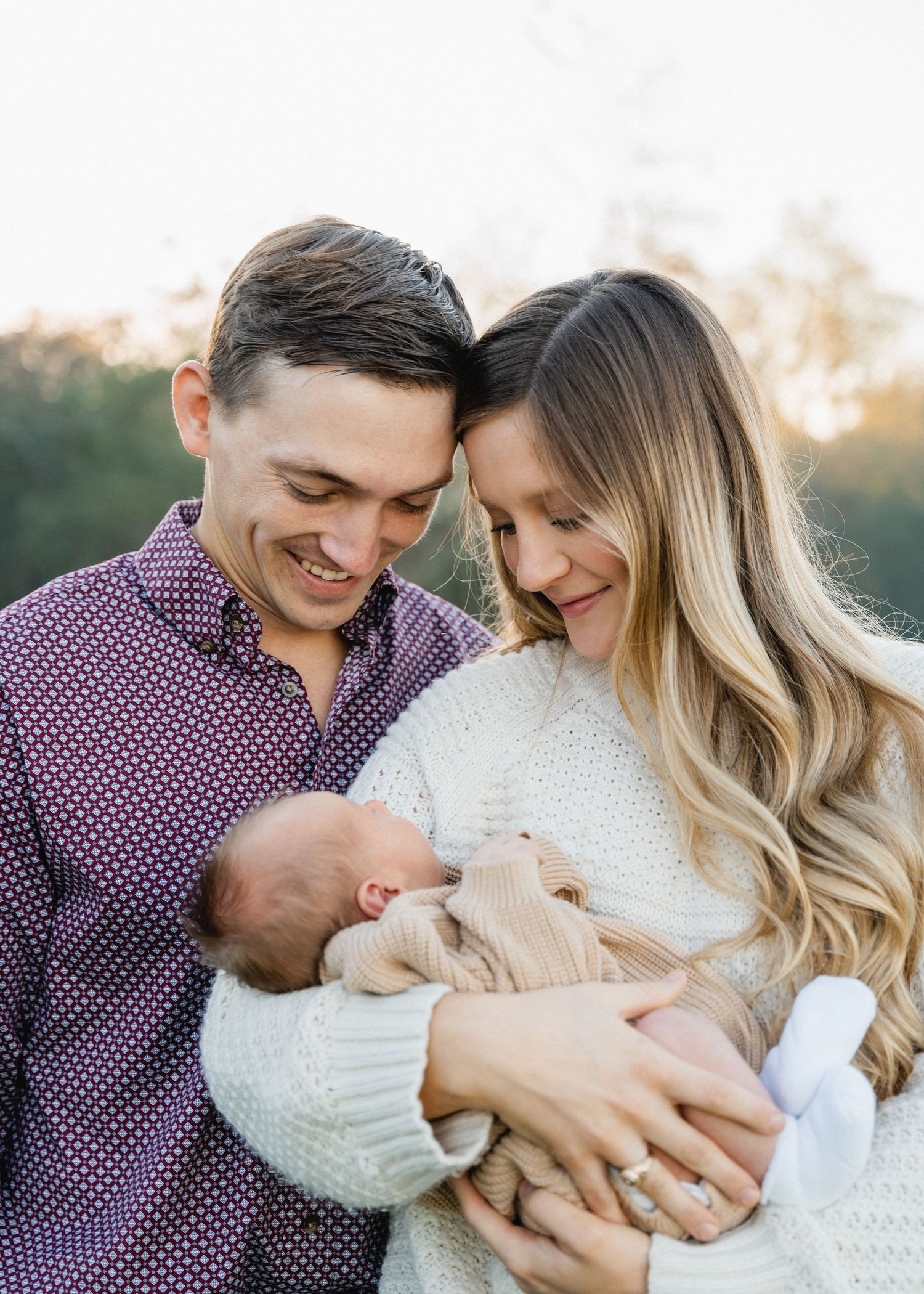 Smiling couple leaning heads together while cradling their newborn baby outdoors at golden hour.