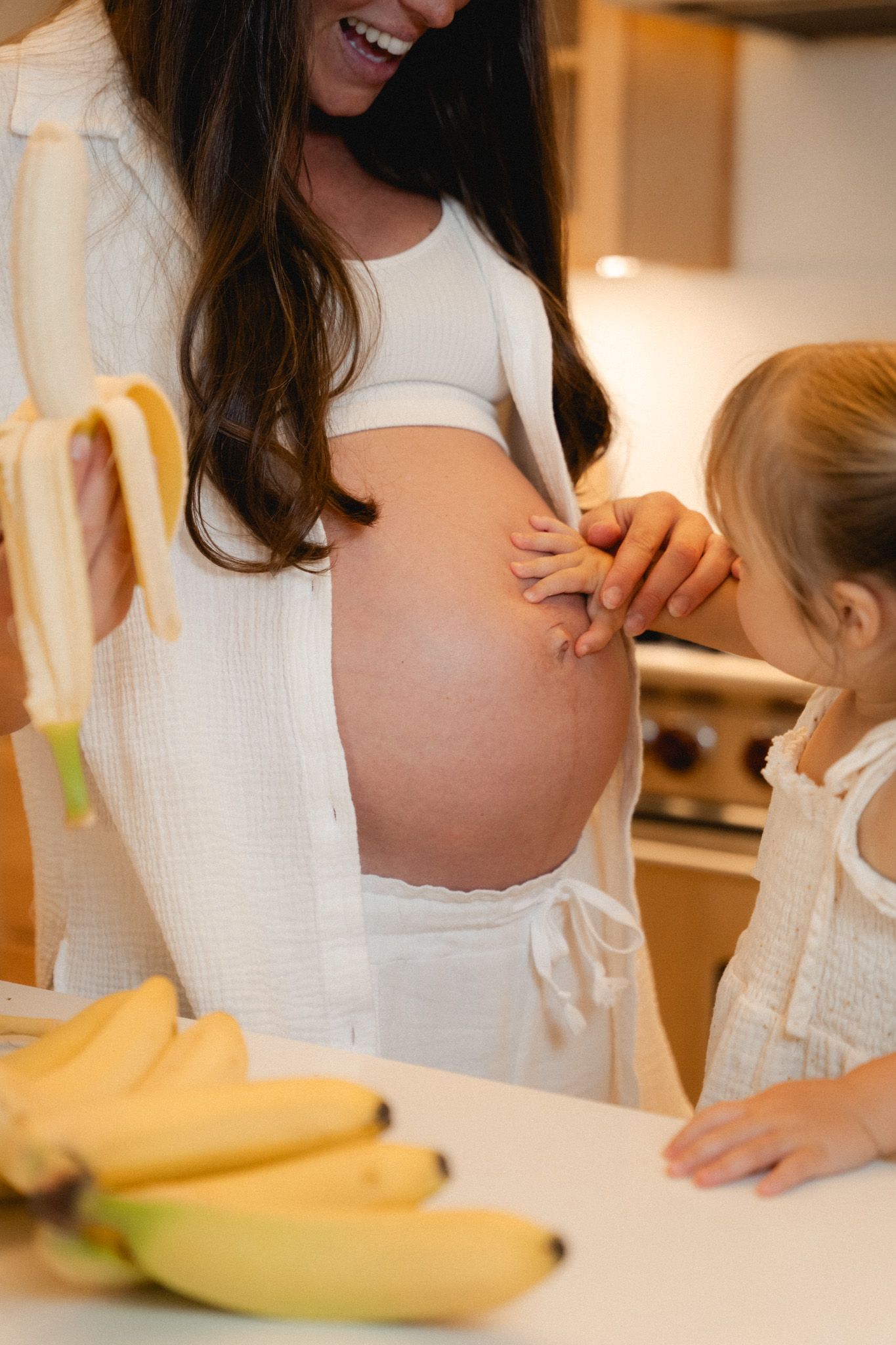 A pregnant woman in white shows her belly to a young girl in a kitchen with bananas on the counter.