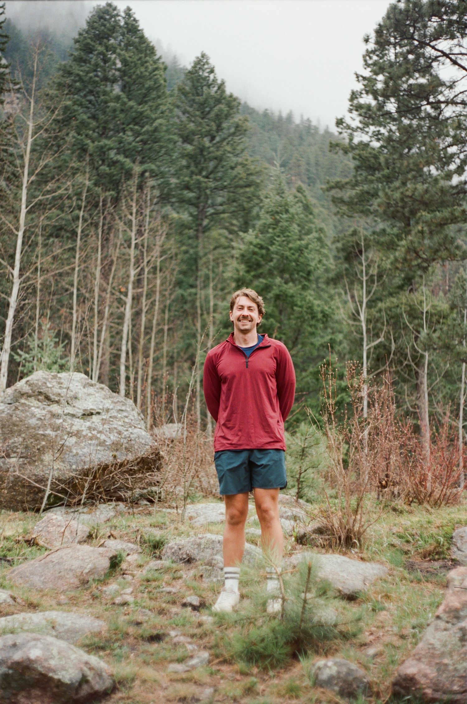 A smiling man in a red hoodie and teal shorts stands on a rocky trail surrounded by tall pine trees in a misty forest.