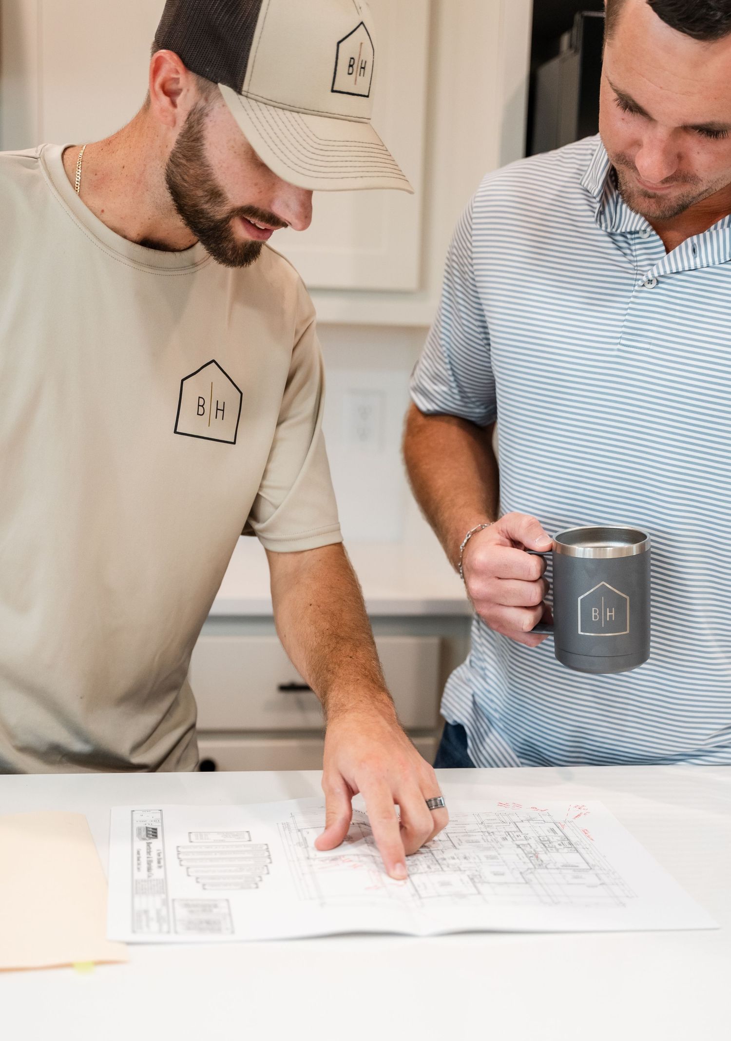 Two men reviewing architectural blueprints on white table holding branded coffee mug.