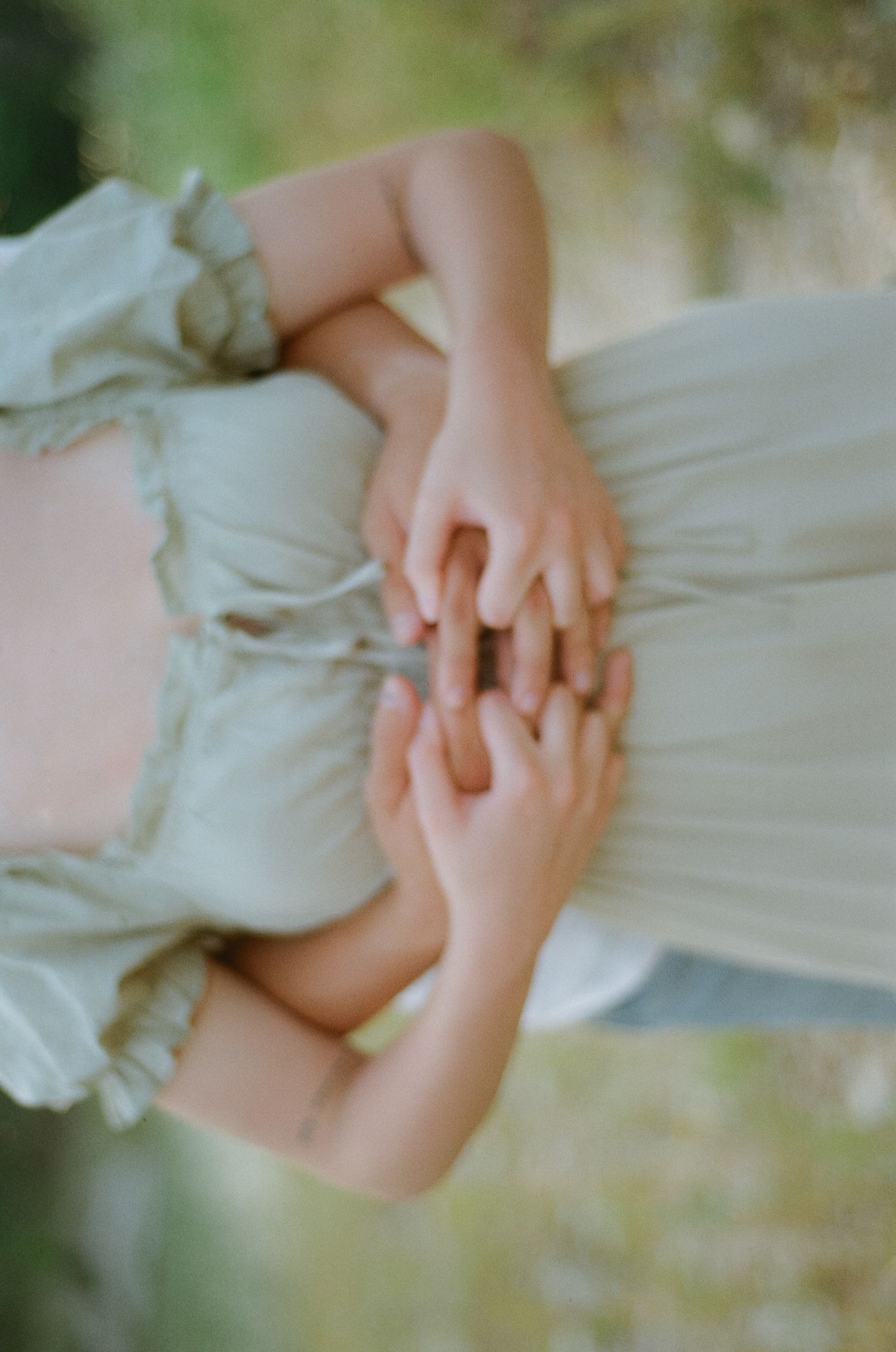 Two people holding hands tenderly, wearing a soft sage green dress, with a blurred green nature background.