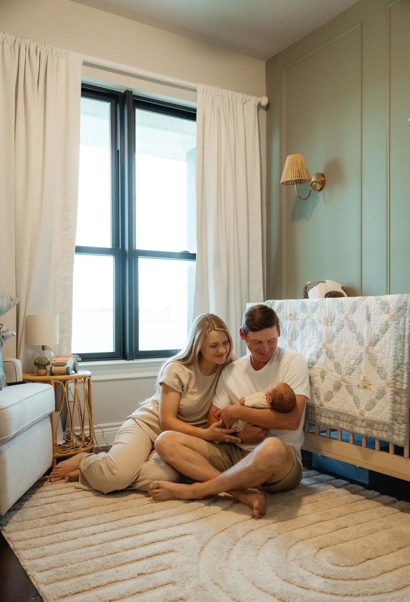 New parents sit on nursery floor holding newborn baby beside a crib in a soft green and neutral toned room.