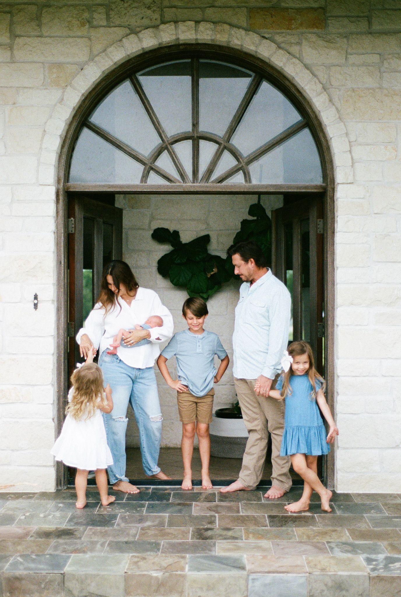 A happy family of six stands barefoot in an elegant arched stone doorway, parents holding their newborn baby.