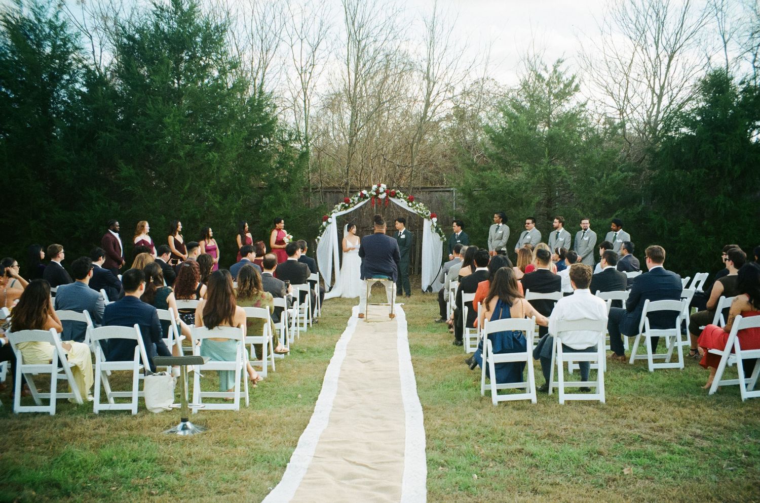 Outdoor wedding ceremony with guests seated in white chairs along a burlap aisle runner.