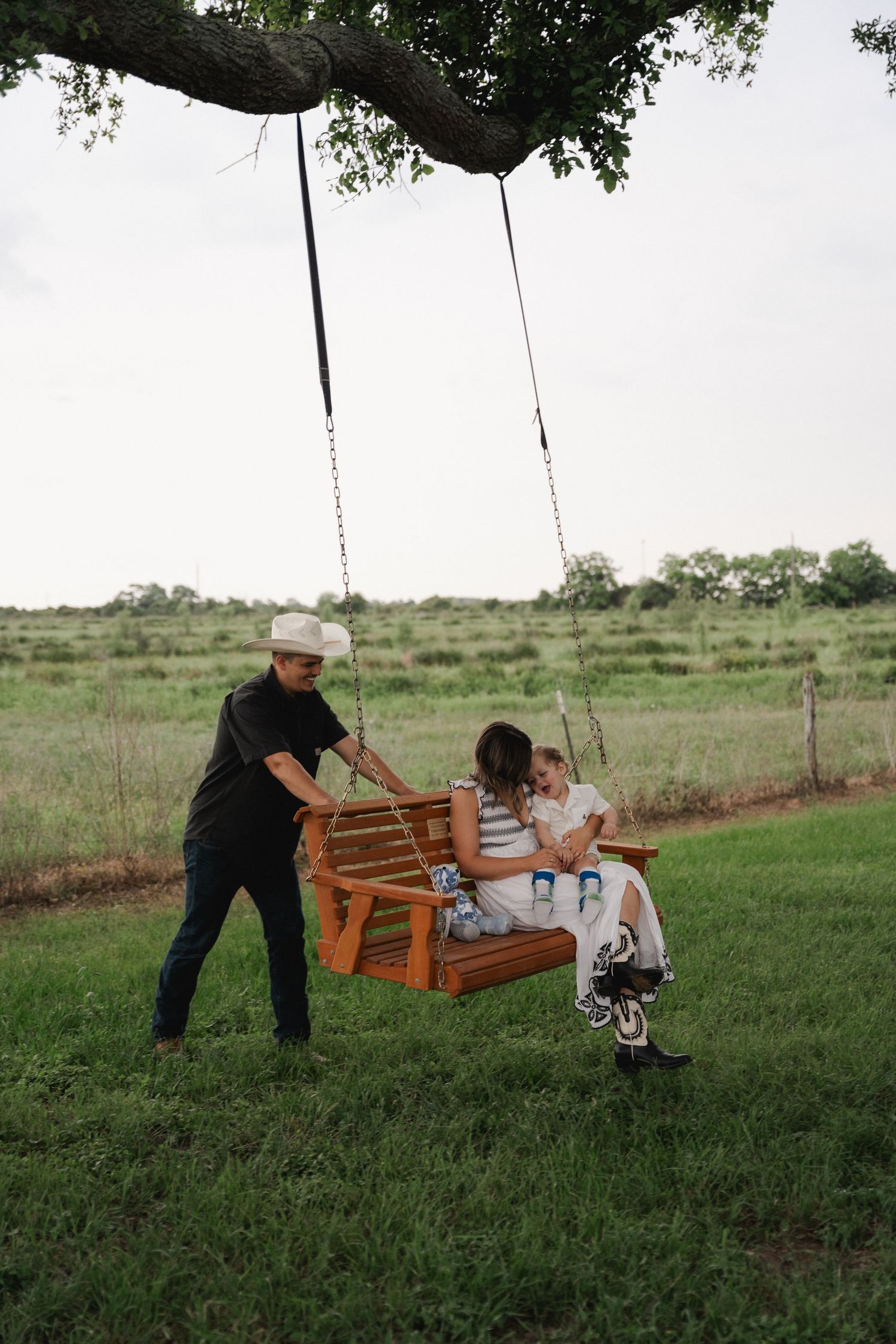 Man pushing woman and toddler on a wooden tree swing in a lush green countryside field.