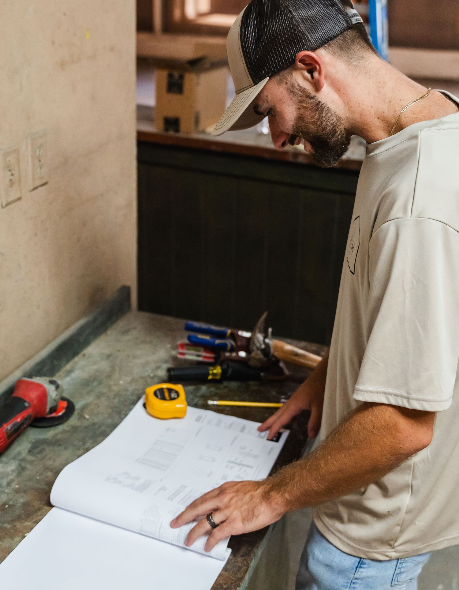 A contractor reviews blueprints on a workbench surrounded by tools including a tape measure and power drill.
