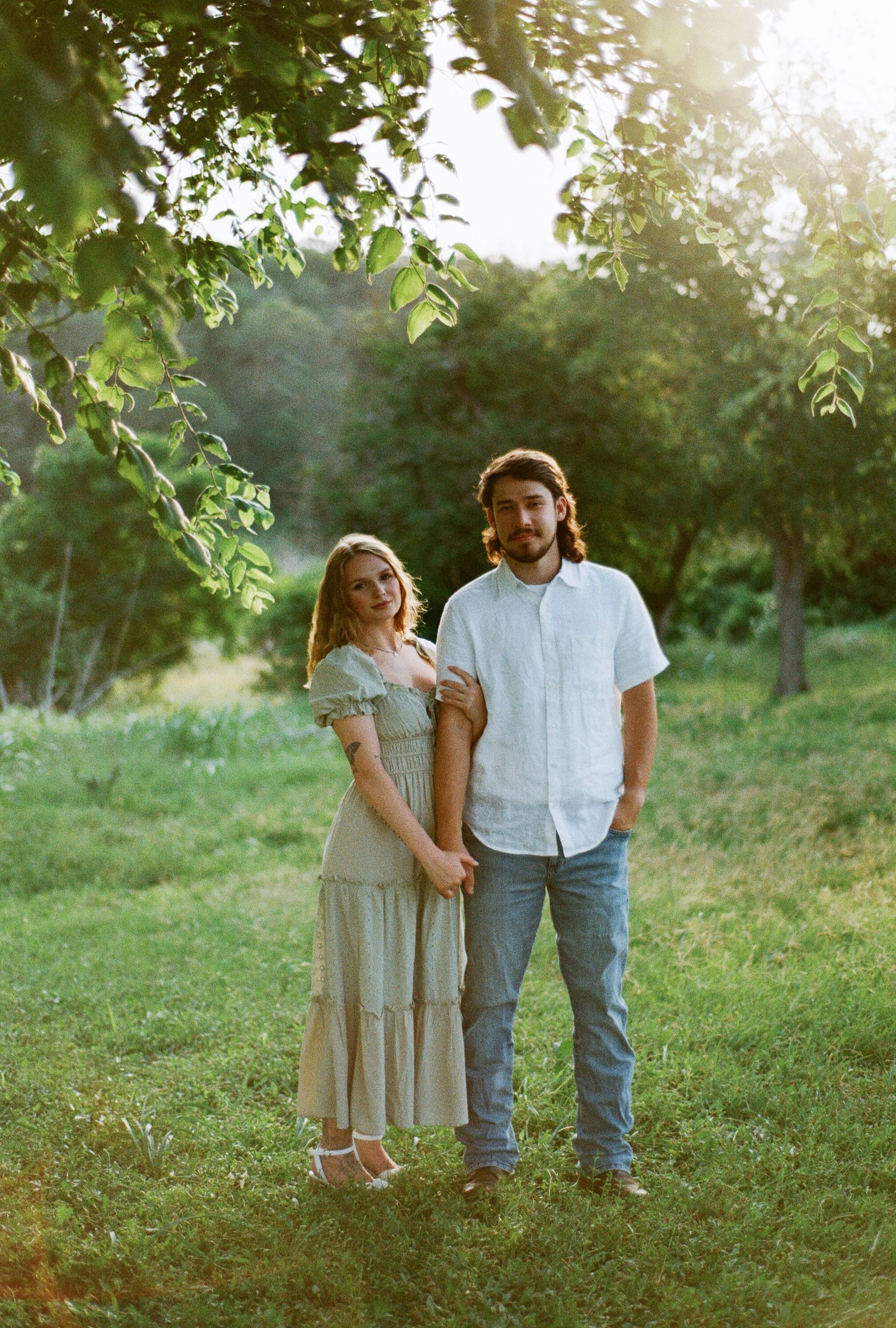 Couple holding hands in a sunlit green meadow, woman in flowy dress and man in white shirt and jeans.