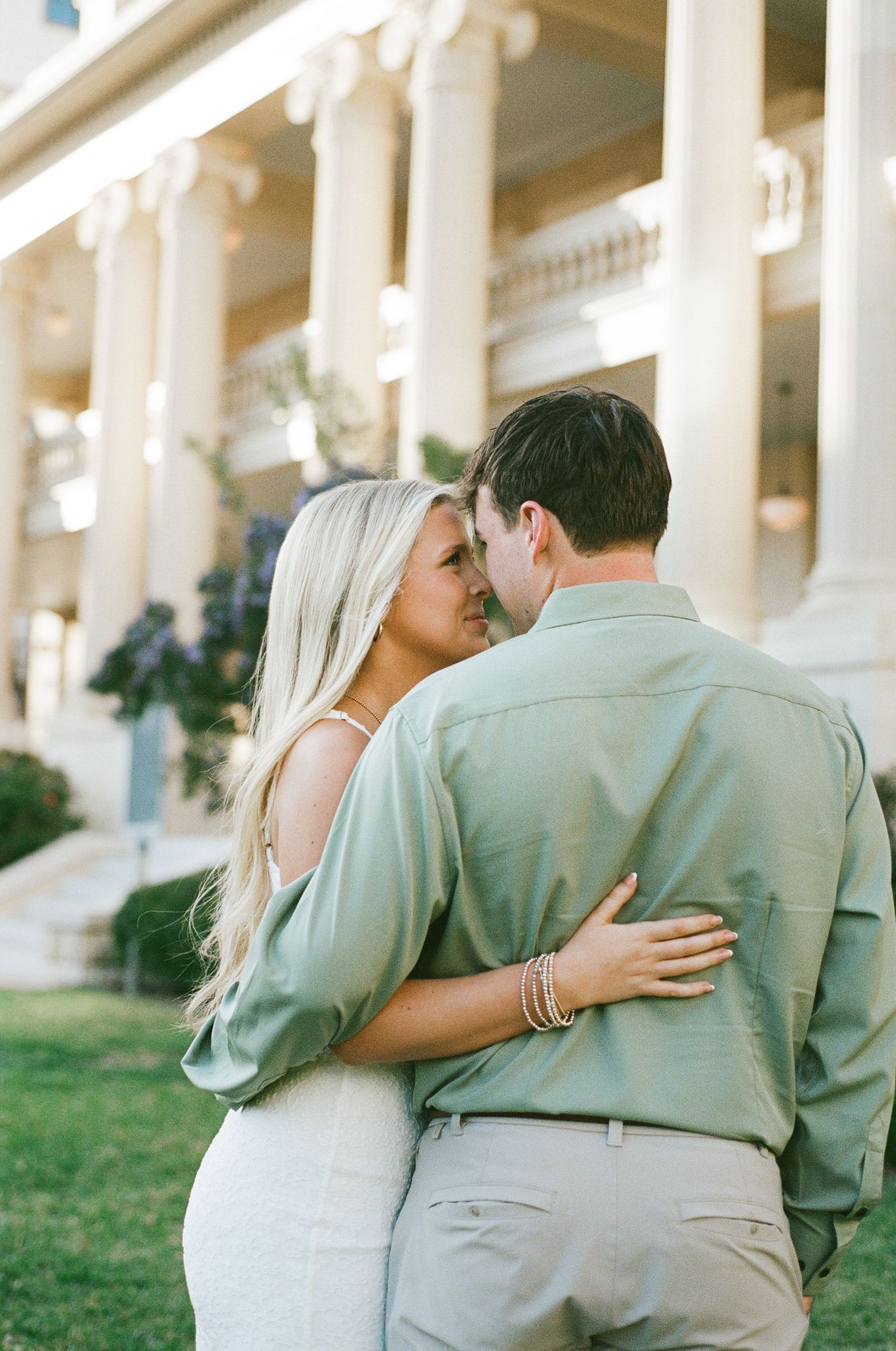 A couple shares a romantic moment embracing near white columns outside an elegant building on a sunny day.