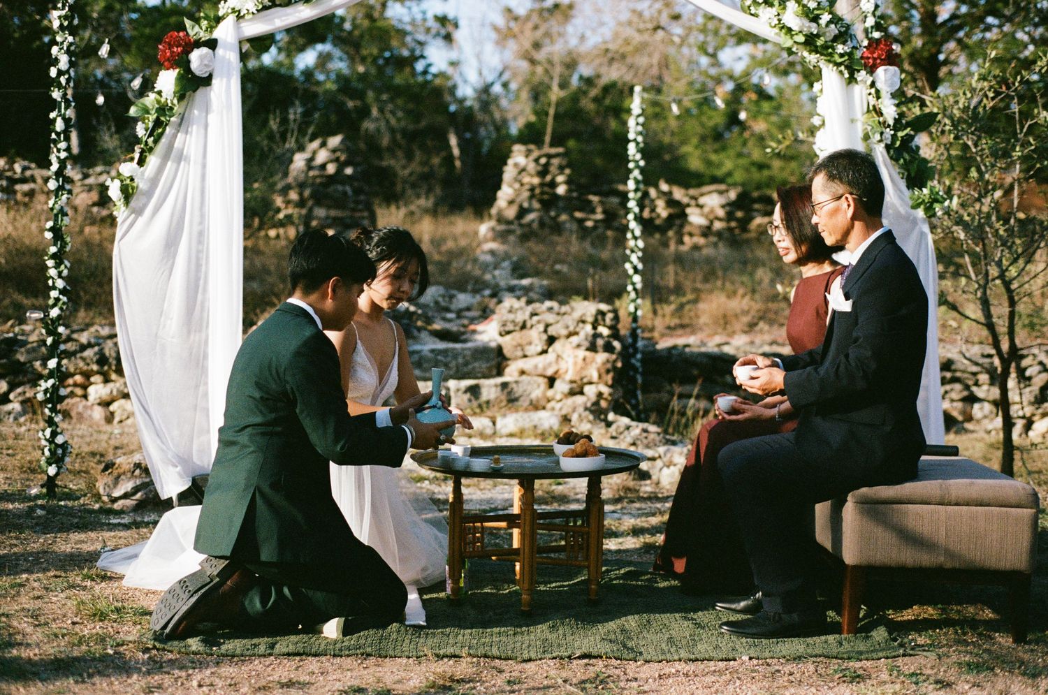 Bride and groom perform traditional Korean tea ceremony with elder at outdoor wedding.