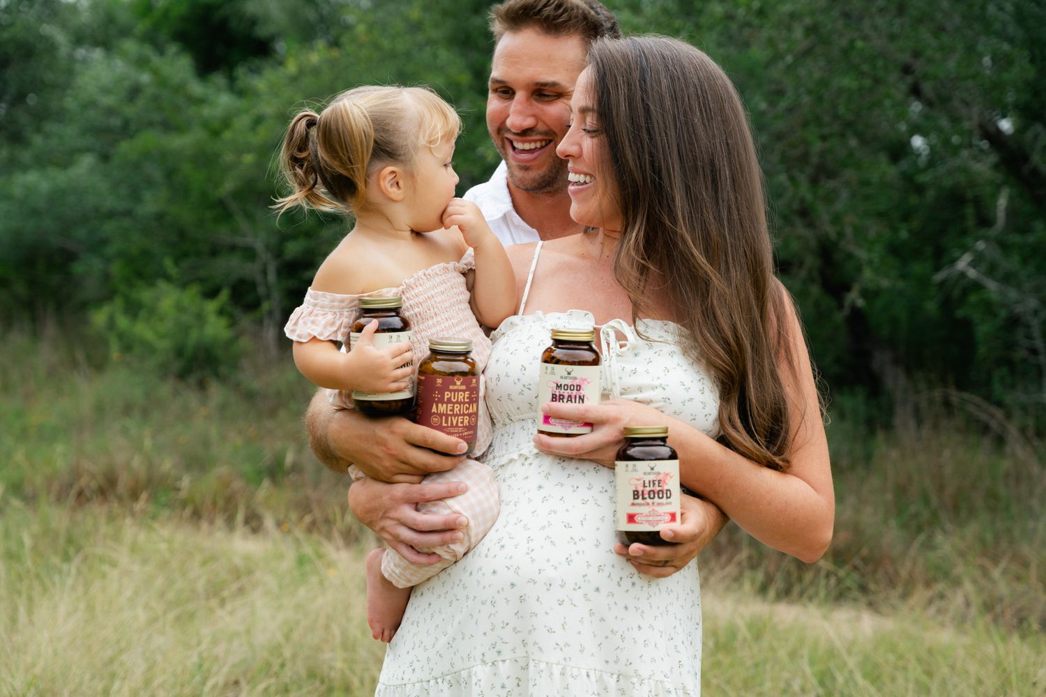 Happy family holding organic food jars outdoors in a grassy field, pregnant woman in white dress with husband and toddler.