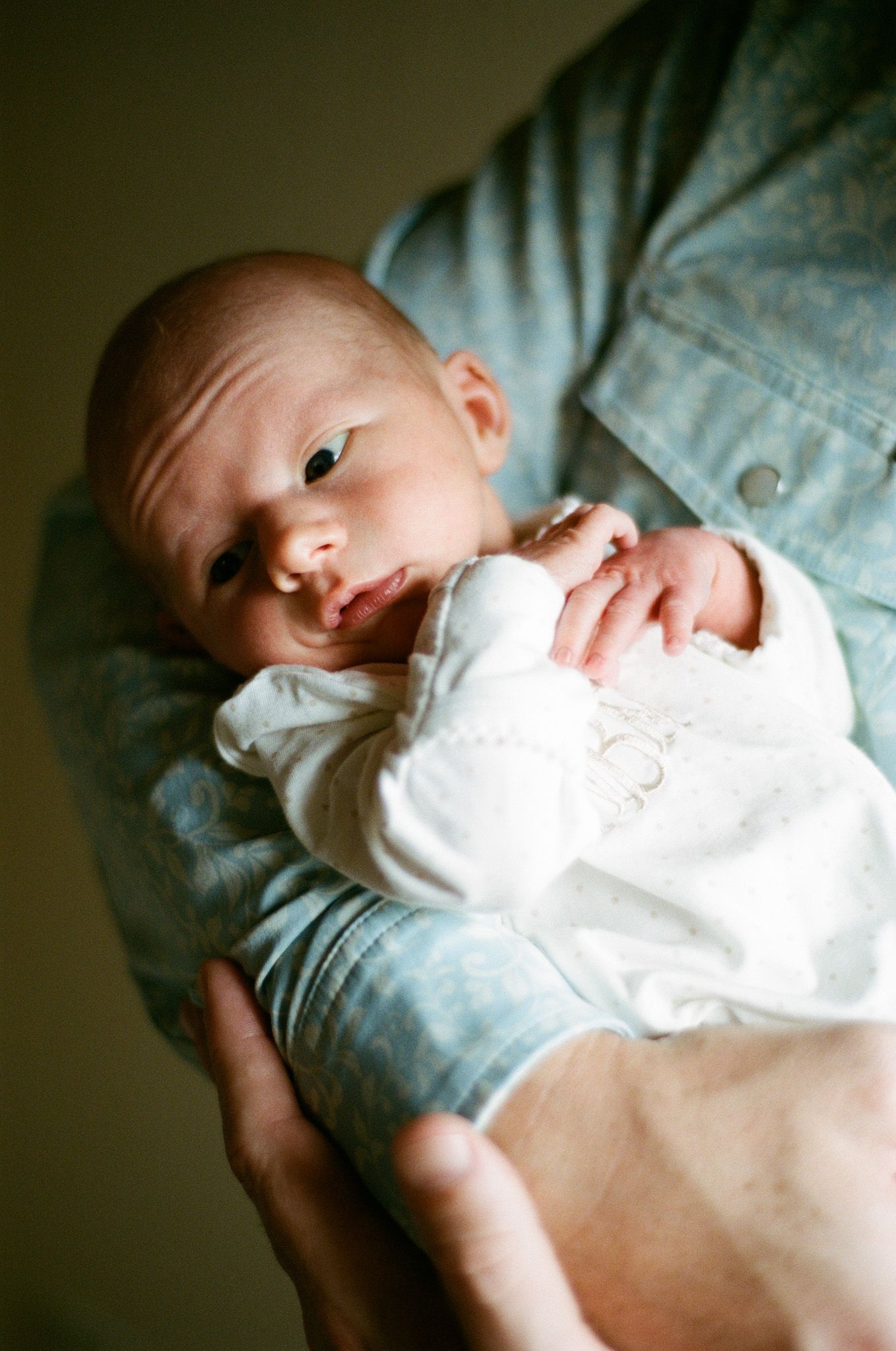 Alert newborn baby in a white outfit being held against an adult's light blue shirt.
