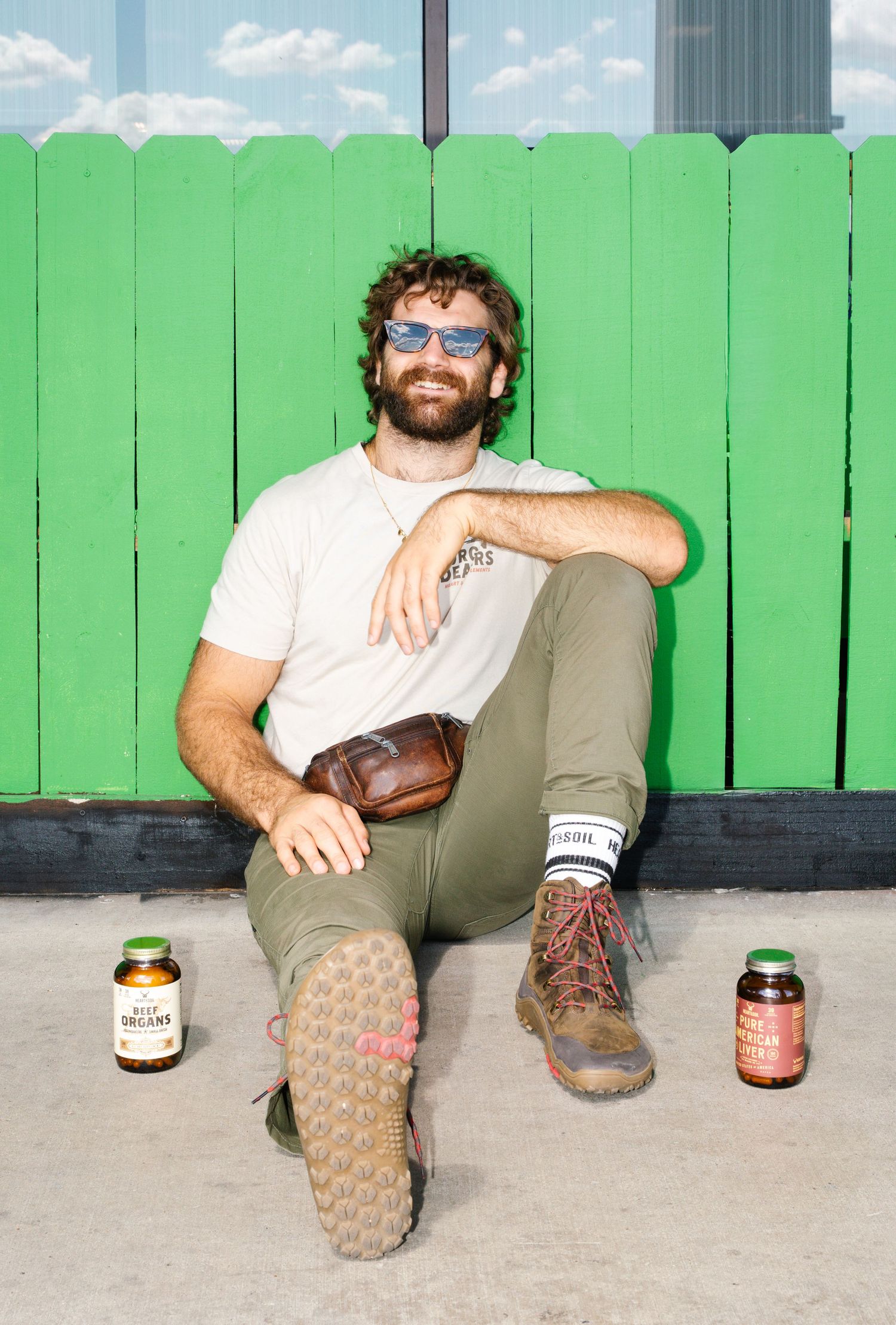 Man in sunglasses and olive pants sits against green fence with supplement bottles beside him.
