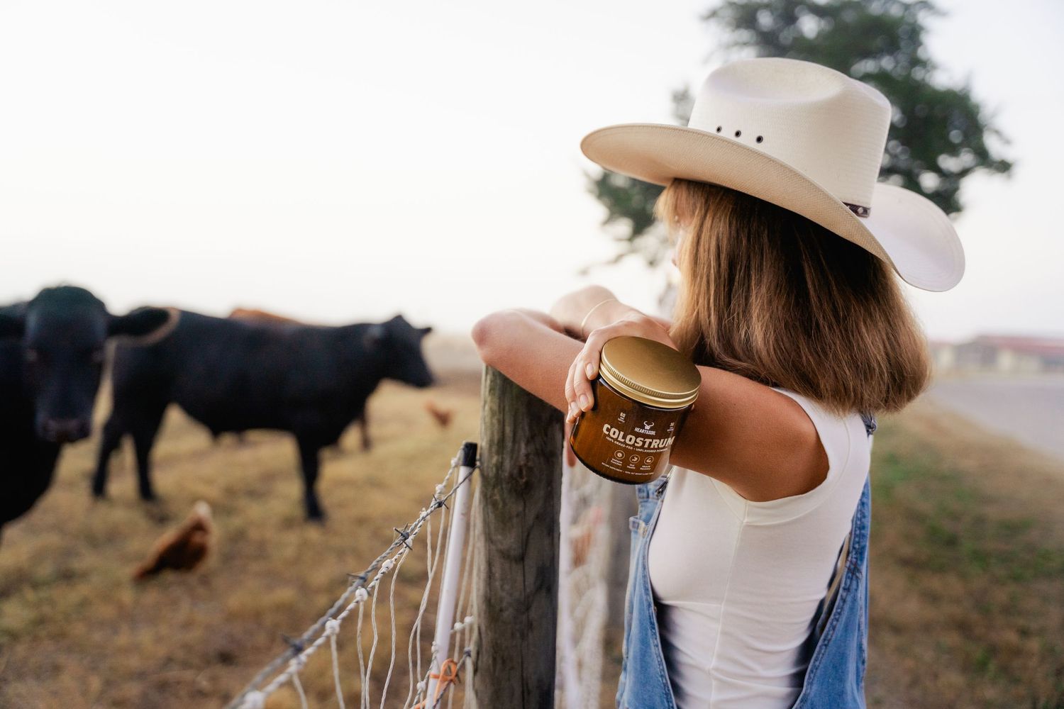 Woman in cowboy hat holding a honey jar leaning on a farm fence with black cattle grazing in background.