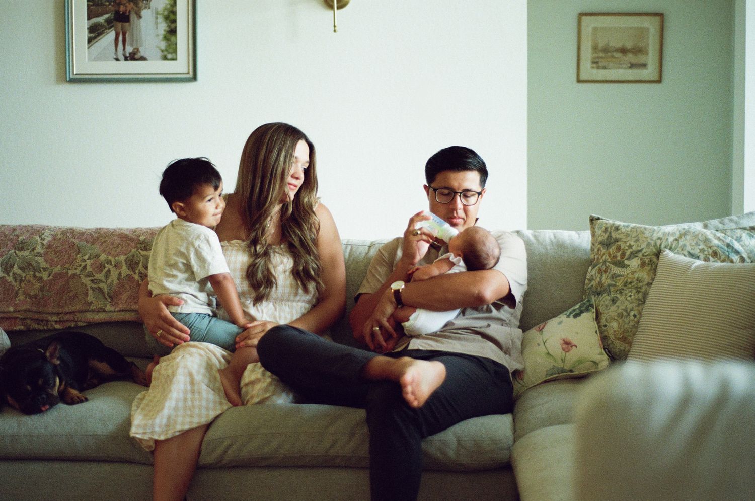 Family of four sitting on a cozy beige sofa, parents with two young children in a warmly decorated living room.