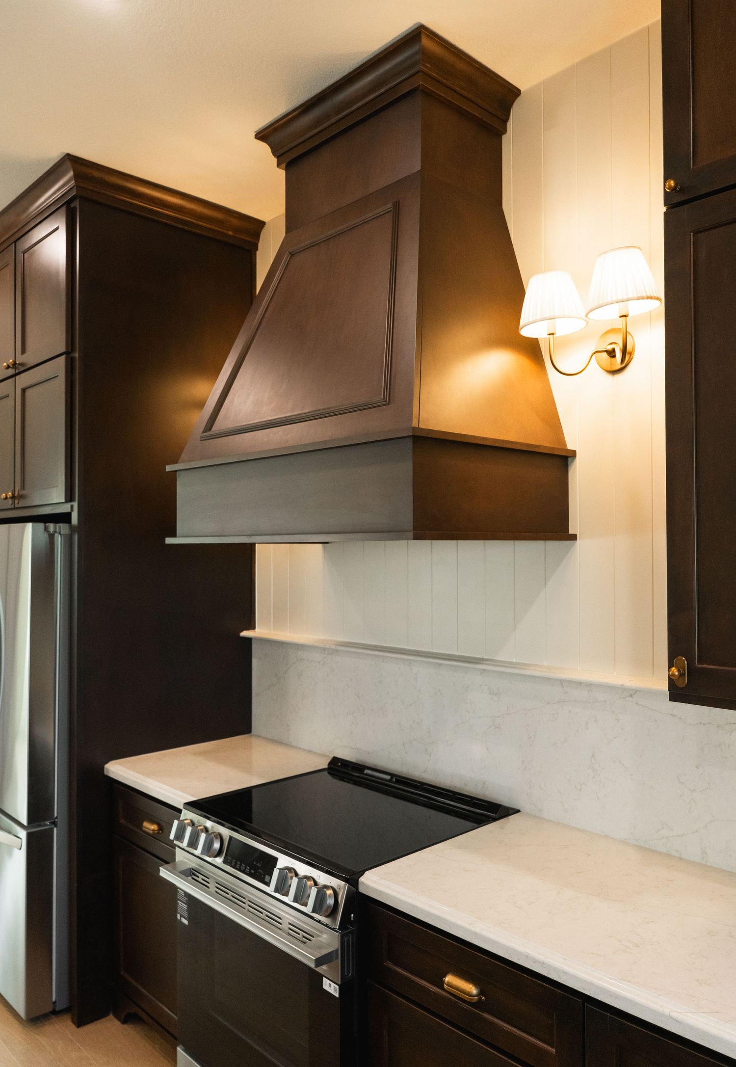 Dark wood kitchen with custom range hood, marble countertops, stainless appliances, and brass sconce.