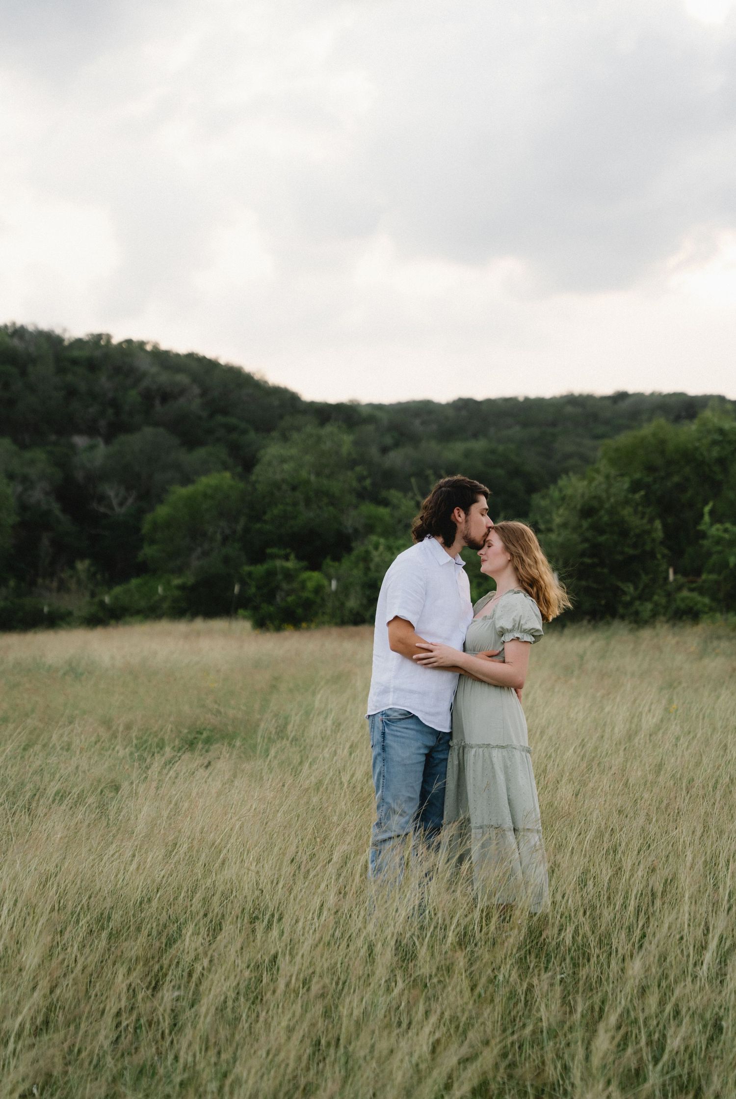 Couple sharing a romantic kiss in a lush green meadow under a cloudy sky in nature.