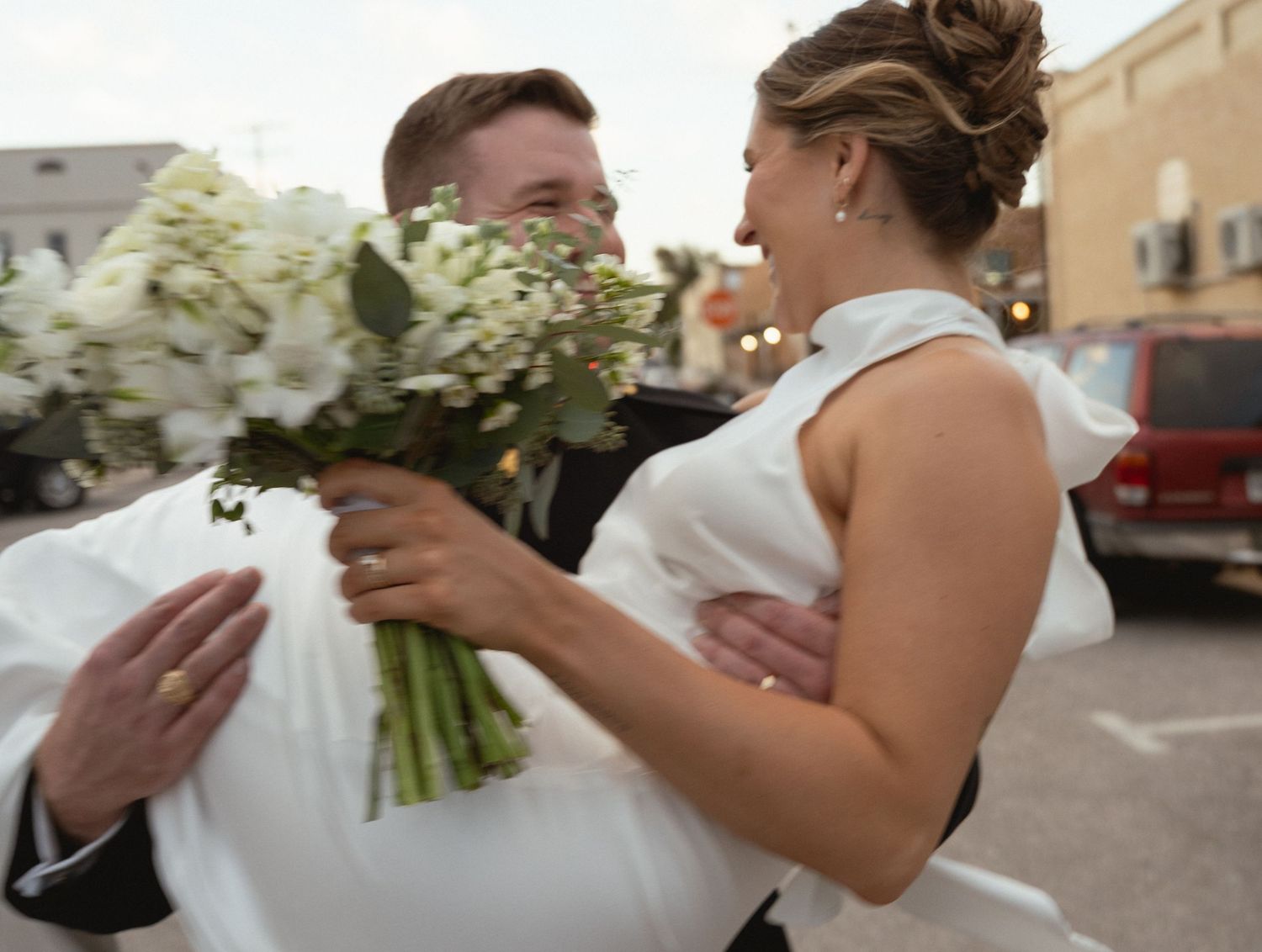 Groom carrying bride in white wedding dress while she holds white flower bouquet outdoors.