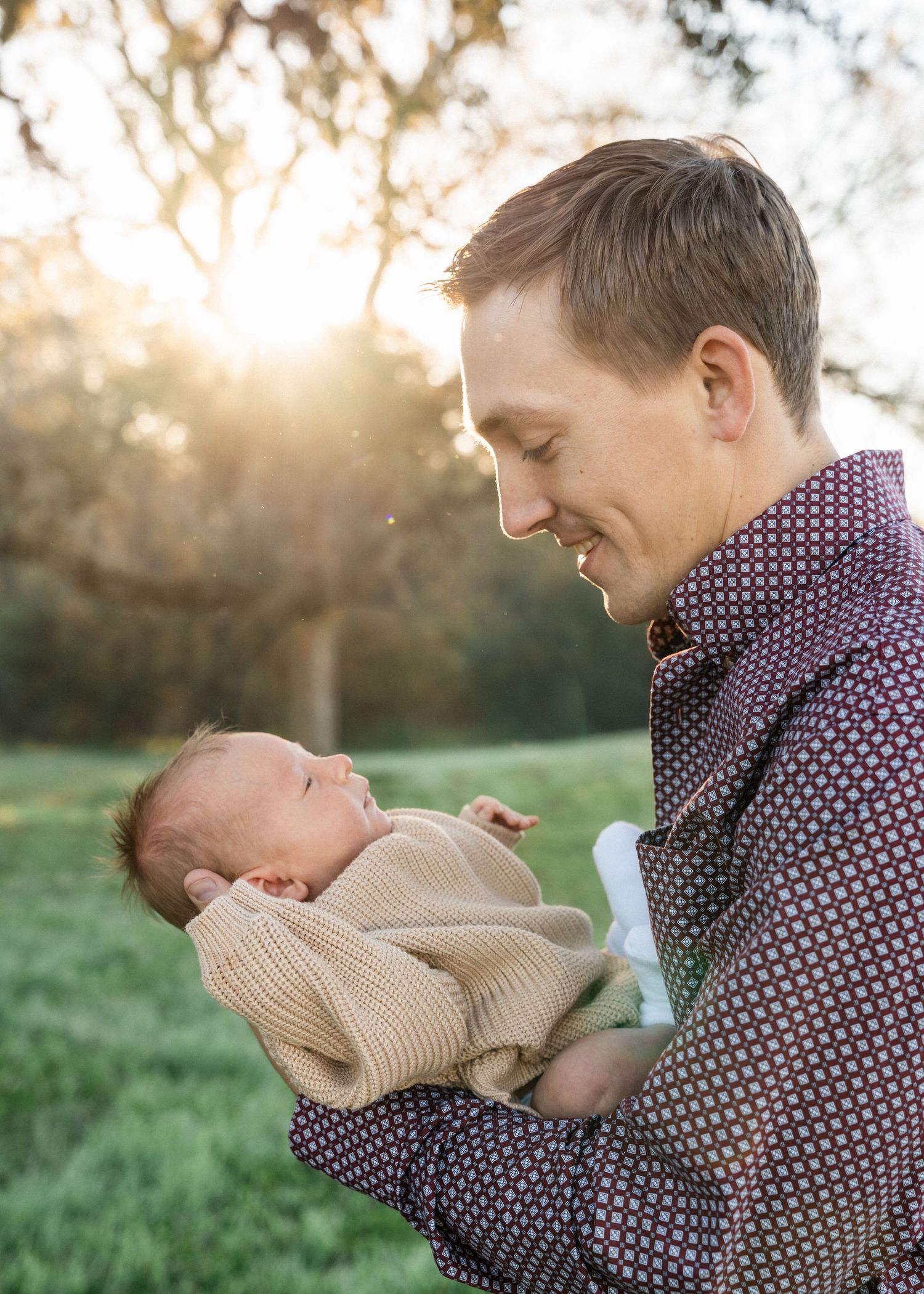 A smiling father cradles his newborn baby outdoors with golden sunlight glowing through trees behind him.