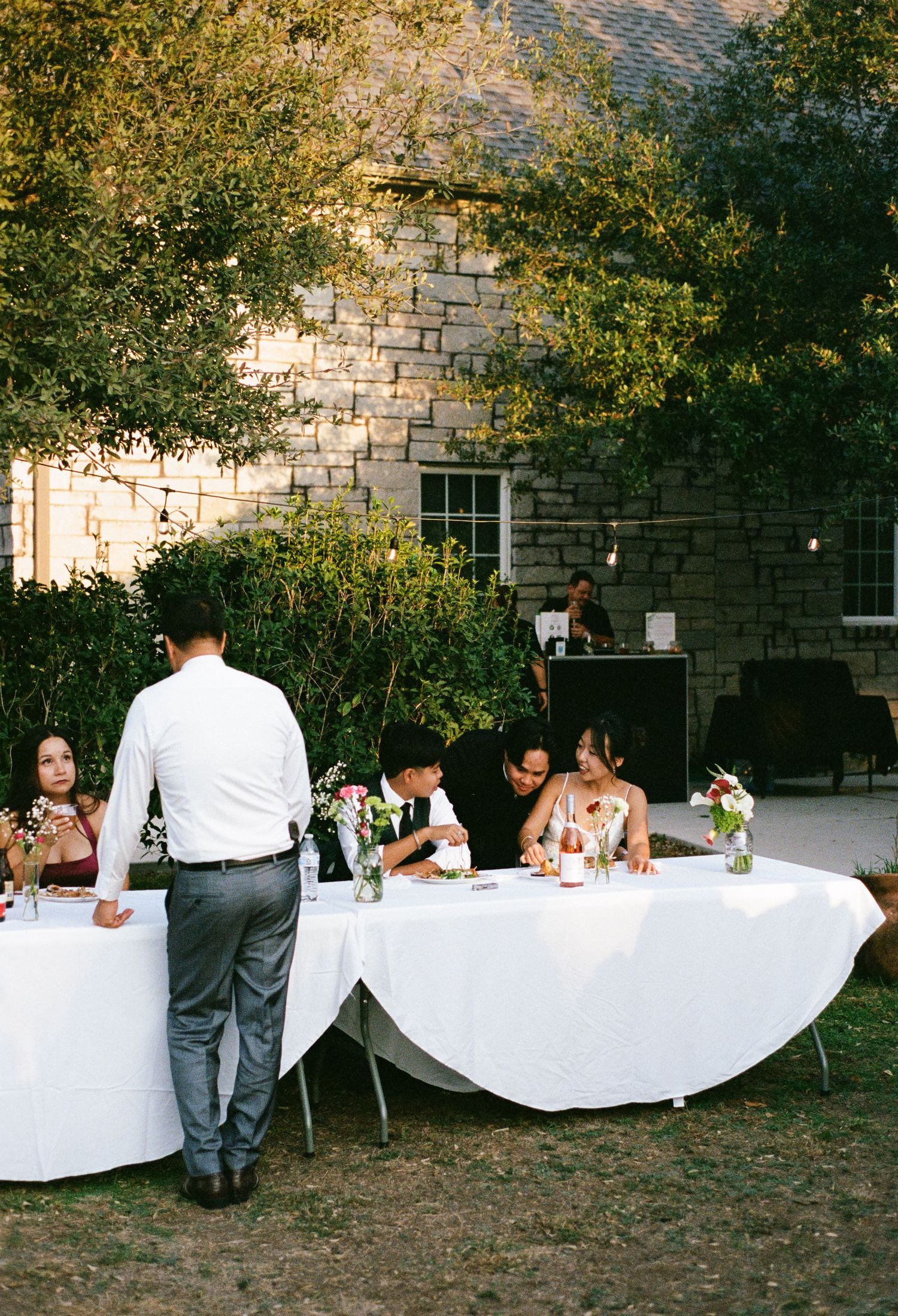 Guests gather around white-clothed tables at an outdoor wedding reception near a stone building.