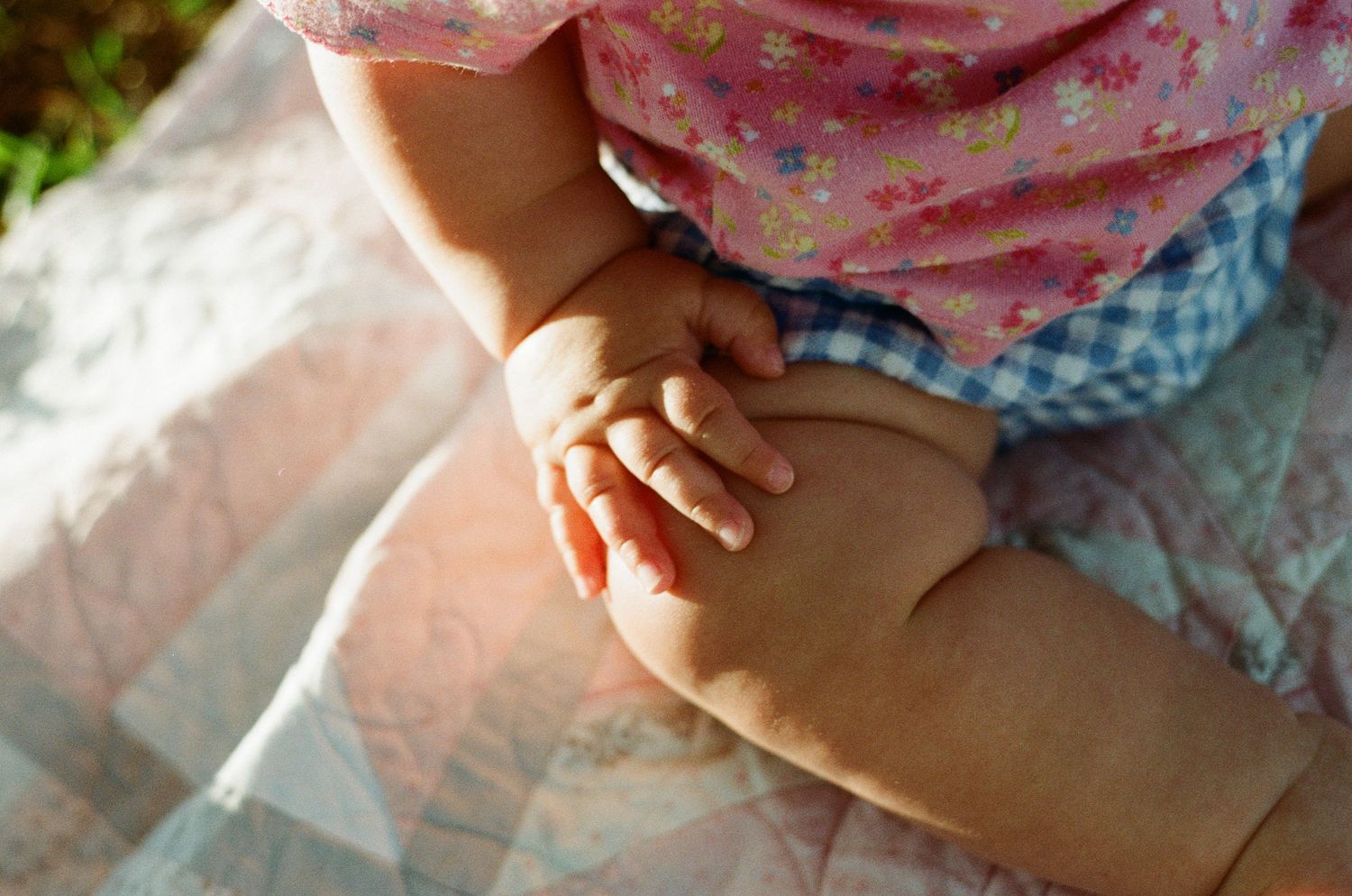Close-up of a baby's chubby hands and legs in a pink floral outfit sitting on a patchwork quilt outdoors.