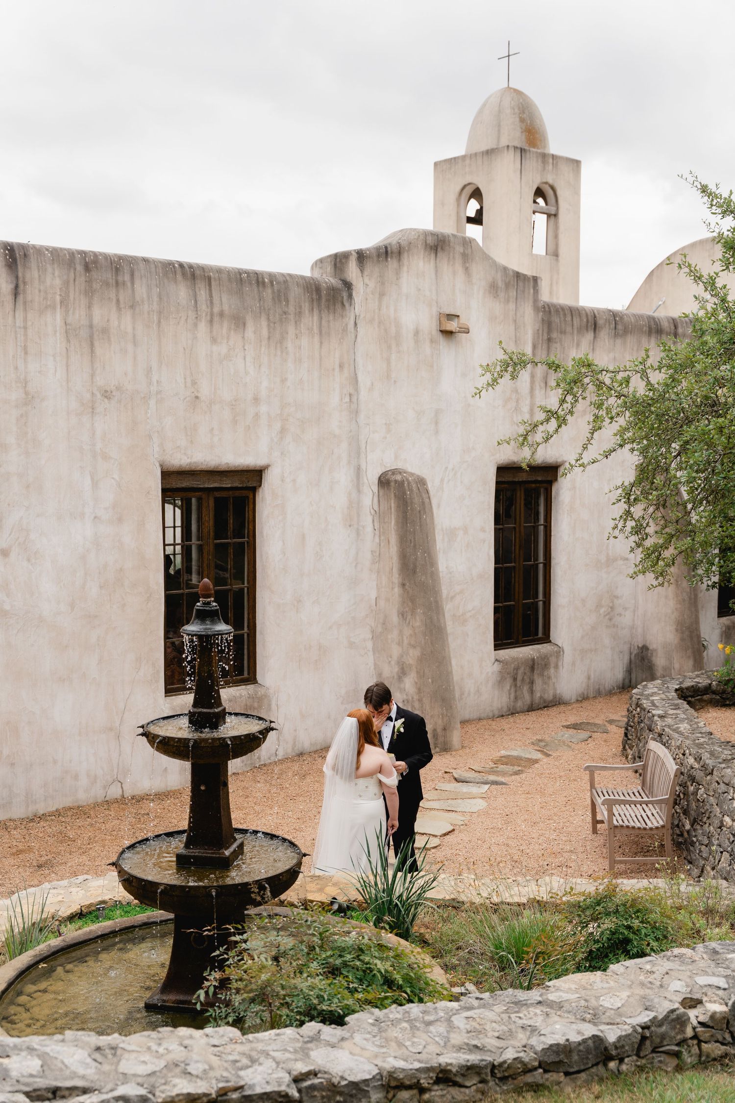 Bride and groom share a romantic embrace in a Spanish mission courtyard with a stone fountain.