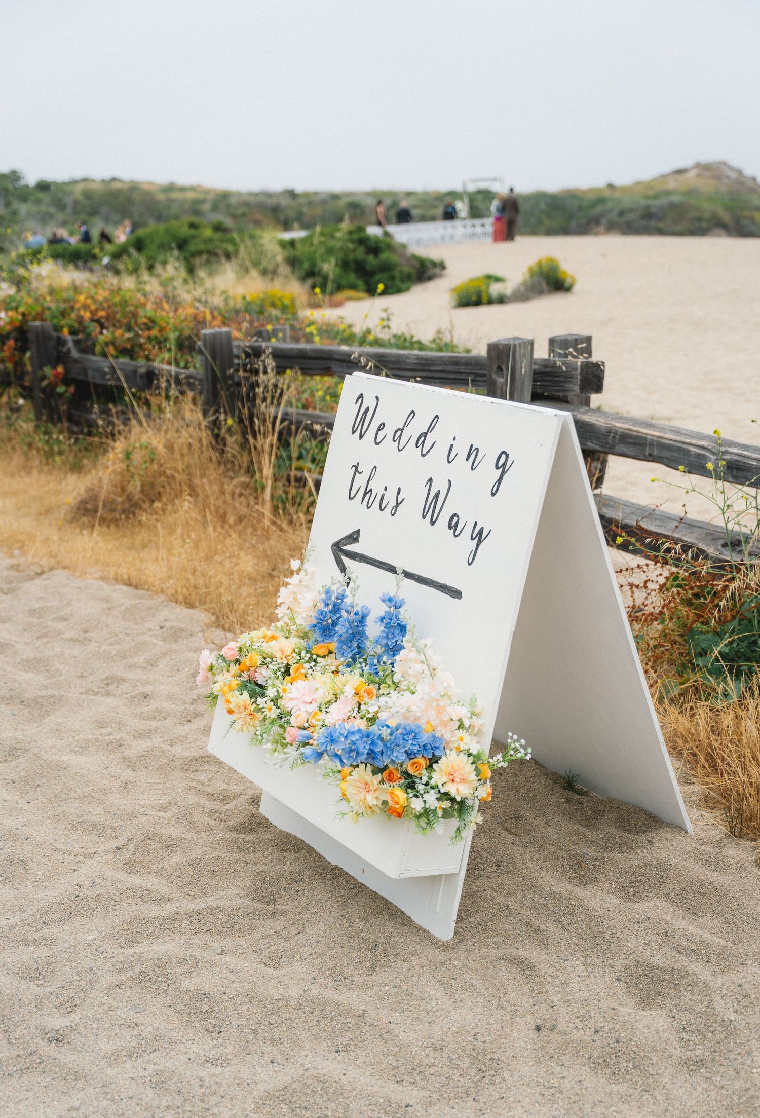 White wedding directional sign with colorful flowers on sandy beach path.