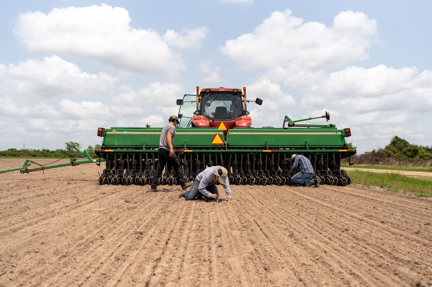 Farmers working alongside a green John Deere tractor planting seeds in a field.