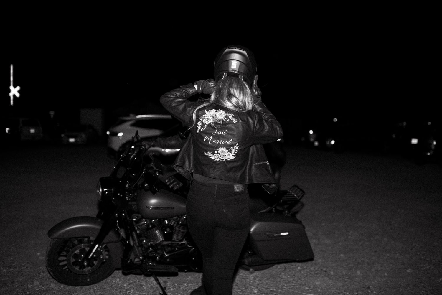 Woman in embroidered leather jacket holding helmet beside a Harley-Davidson motorcycle at night.