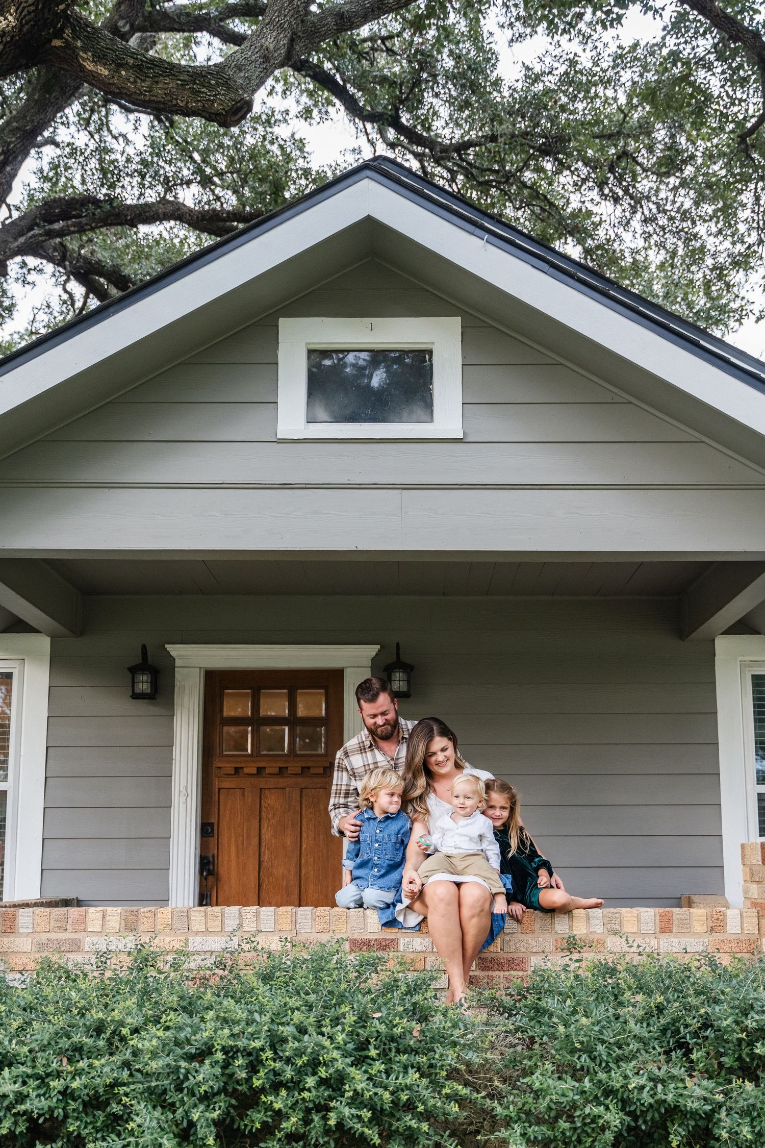 A happy family of four sits together on the front porch steps of a charming gray cottage home.