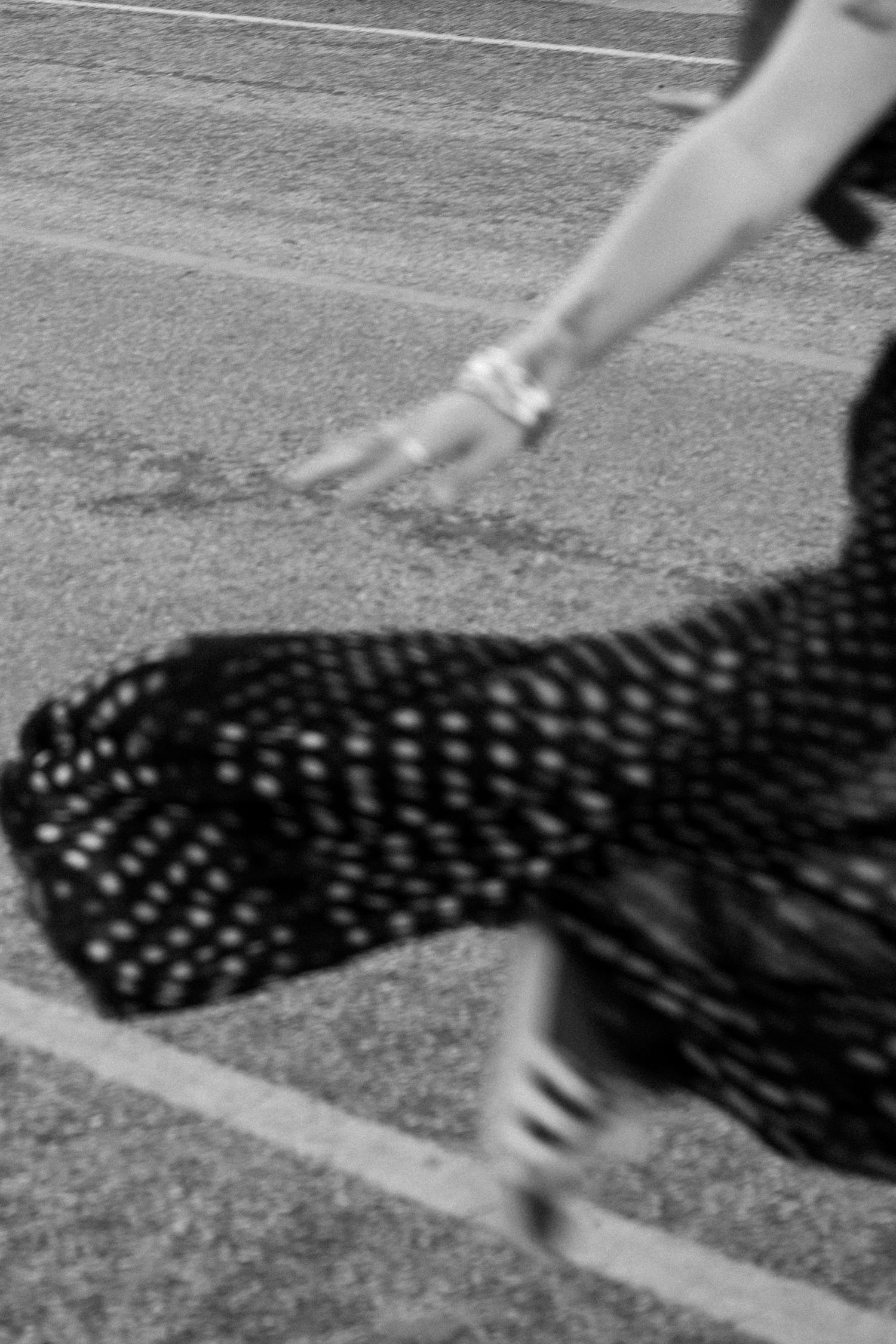 Black and white motion blur photo of woman in polka dot skirt walking on pavement street.