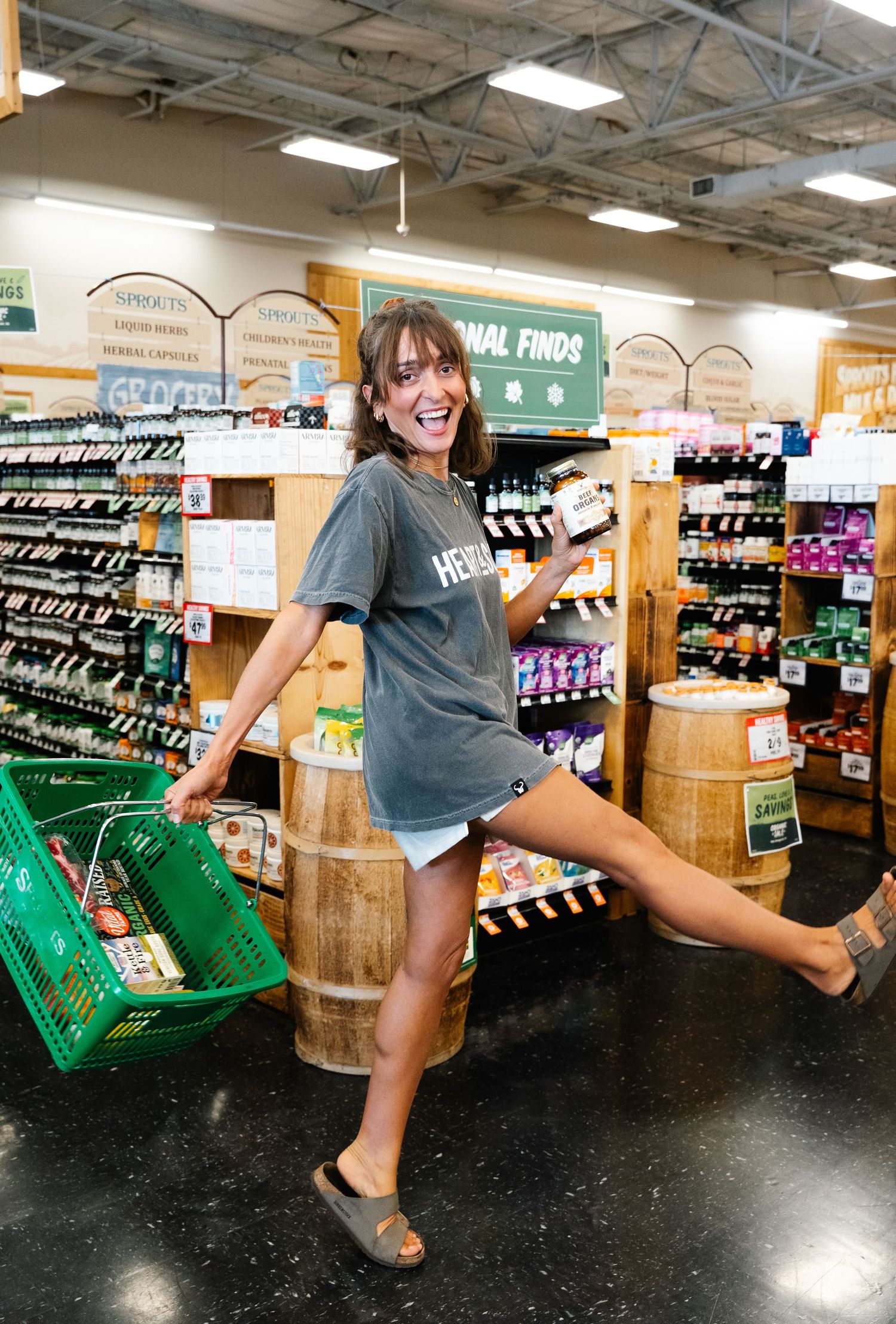 Woman in gray t-shirt poses playfully with green basket in a natural foods grocery store aisle.