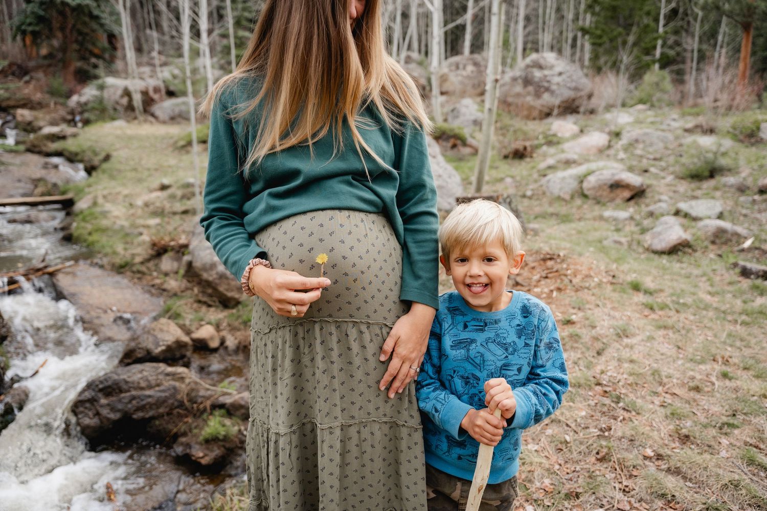 Pregnant woman in green dress stands outdoors with smiling toddler boy holding stick near rocky stream.