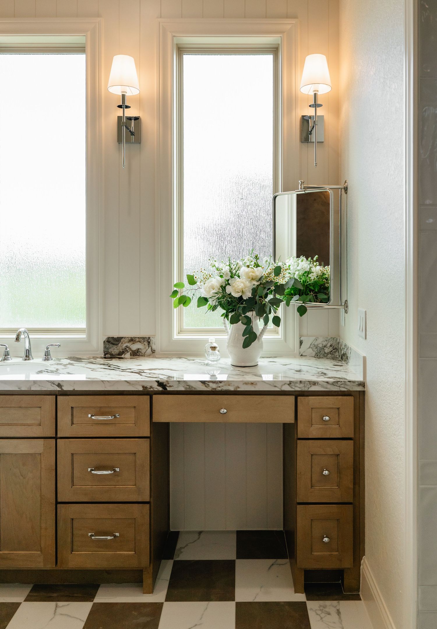 Elegant bathroom vanity with marble countertop, wood cabinets, checkered floors, and floral arrangement.