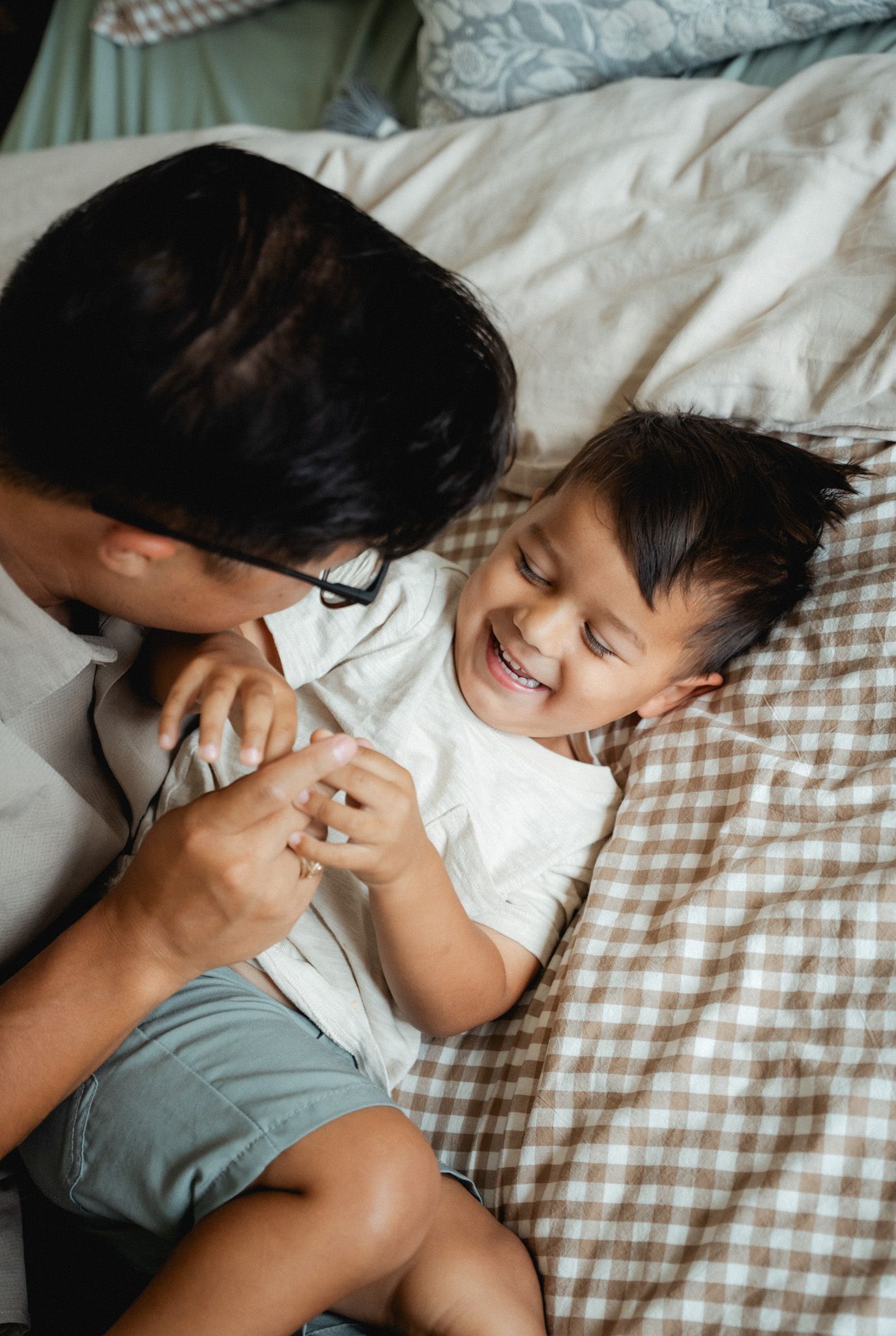A joyful toddler laughs while lying on a plaid bed as an adult playfully interacts with them indoors.