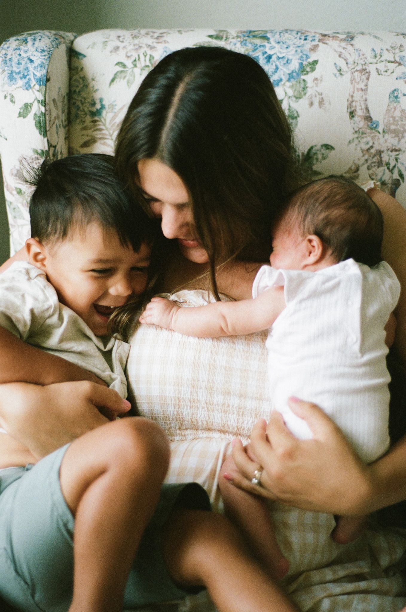 A mother sits in a floral armchair, smiling at her laughing toddler while cradling her newborn baby in her arms.